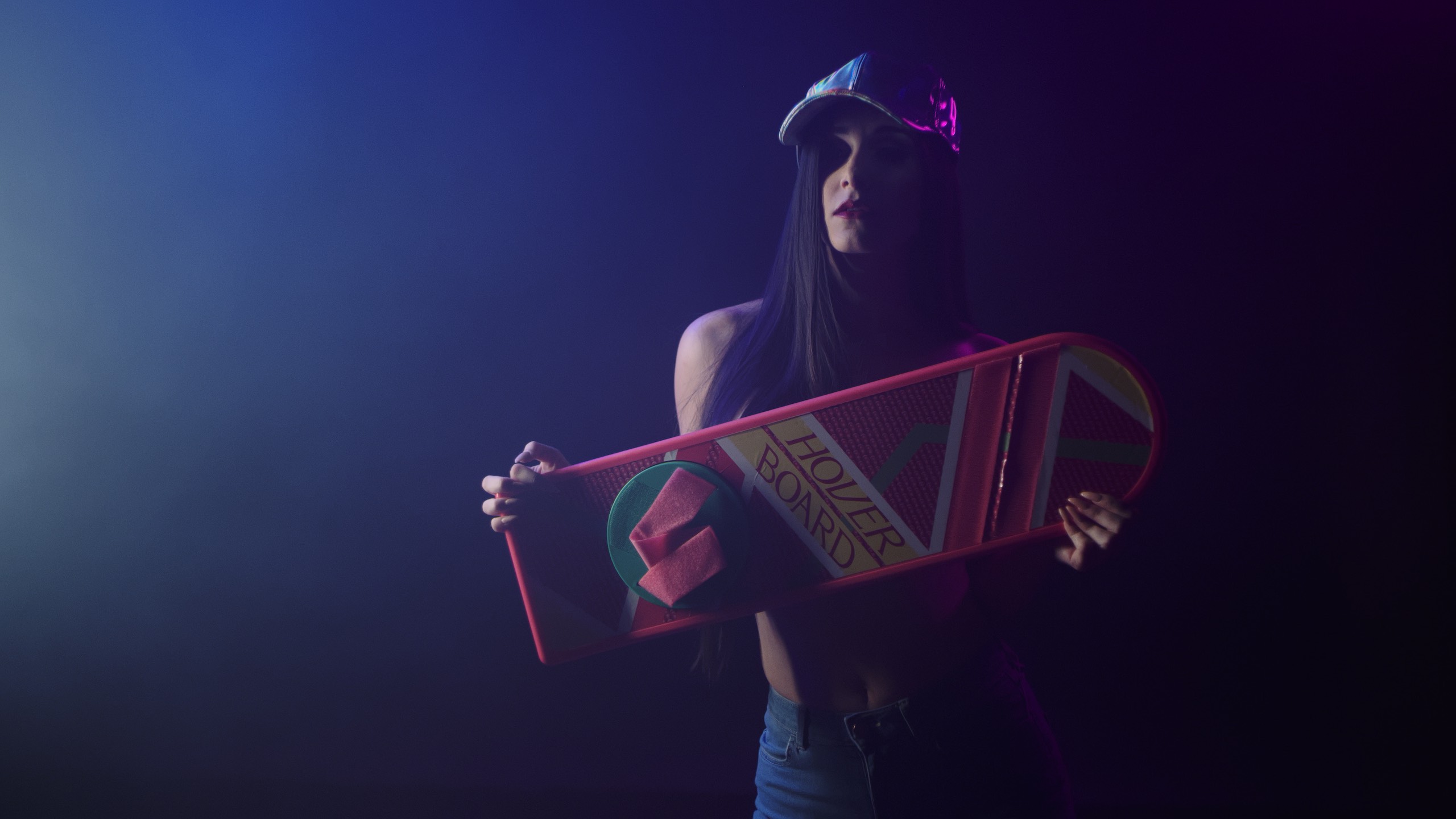 The Delorean Uncreative Music Woman holding a hover board and wearing a multicolored cap and jeans in semi darkness