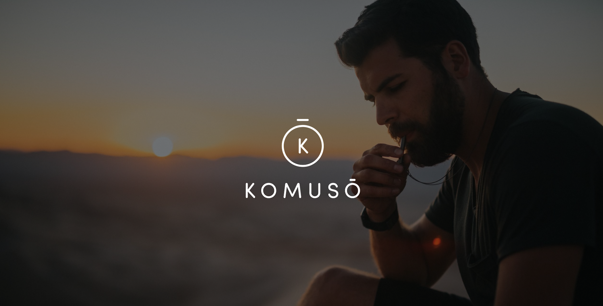 White Komuso logo with dimmed background of a bearded man blowing a whistle with a view of the sun setting in the valley behind him