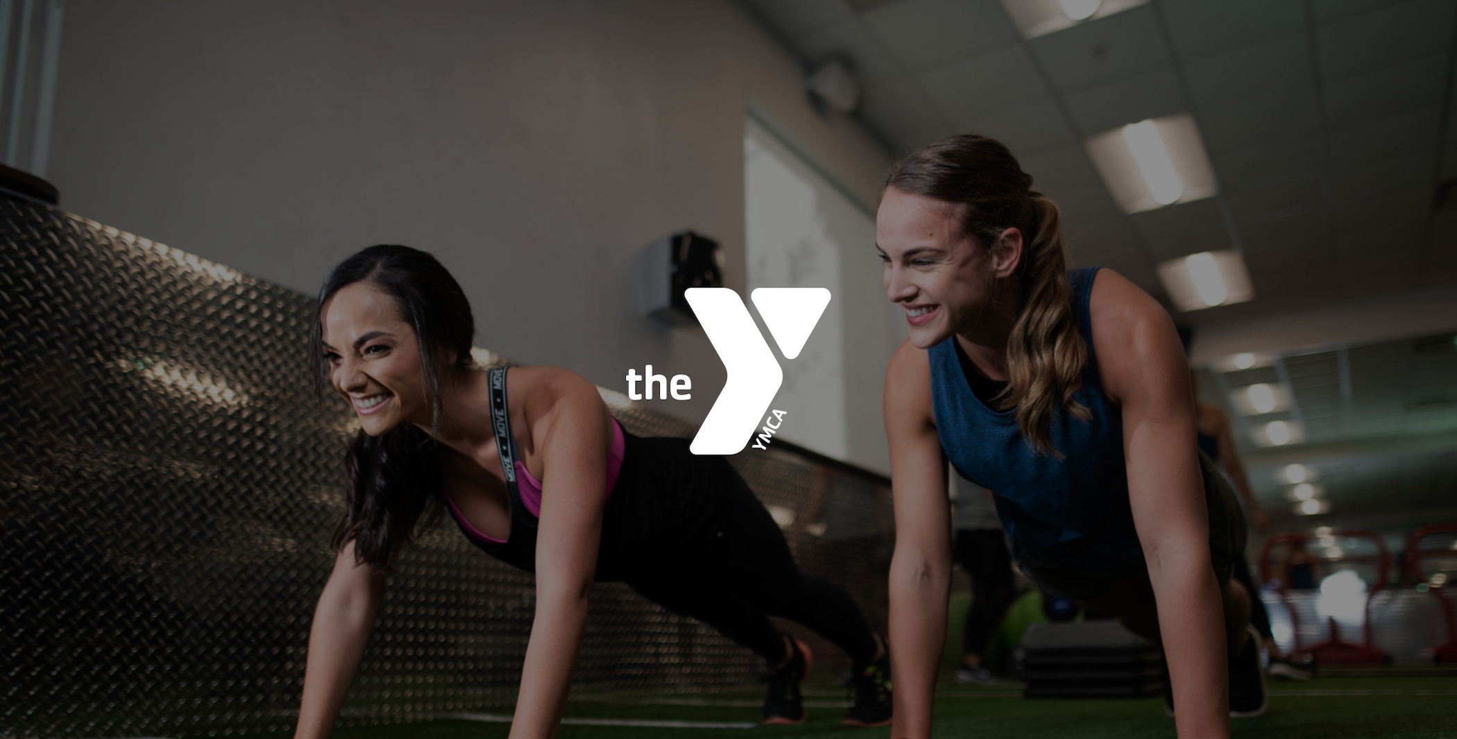 White The YMCA logo with two smiling women in plank poses in a gym