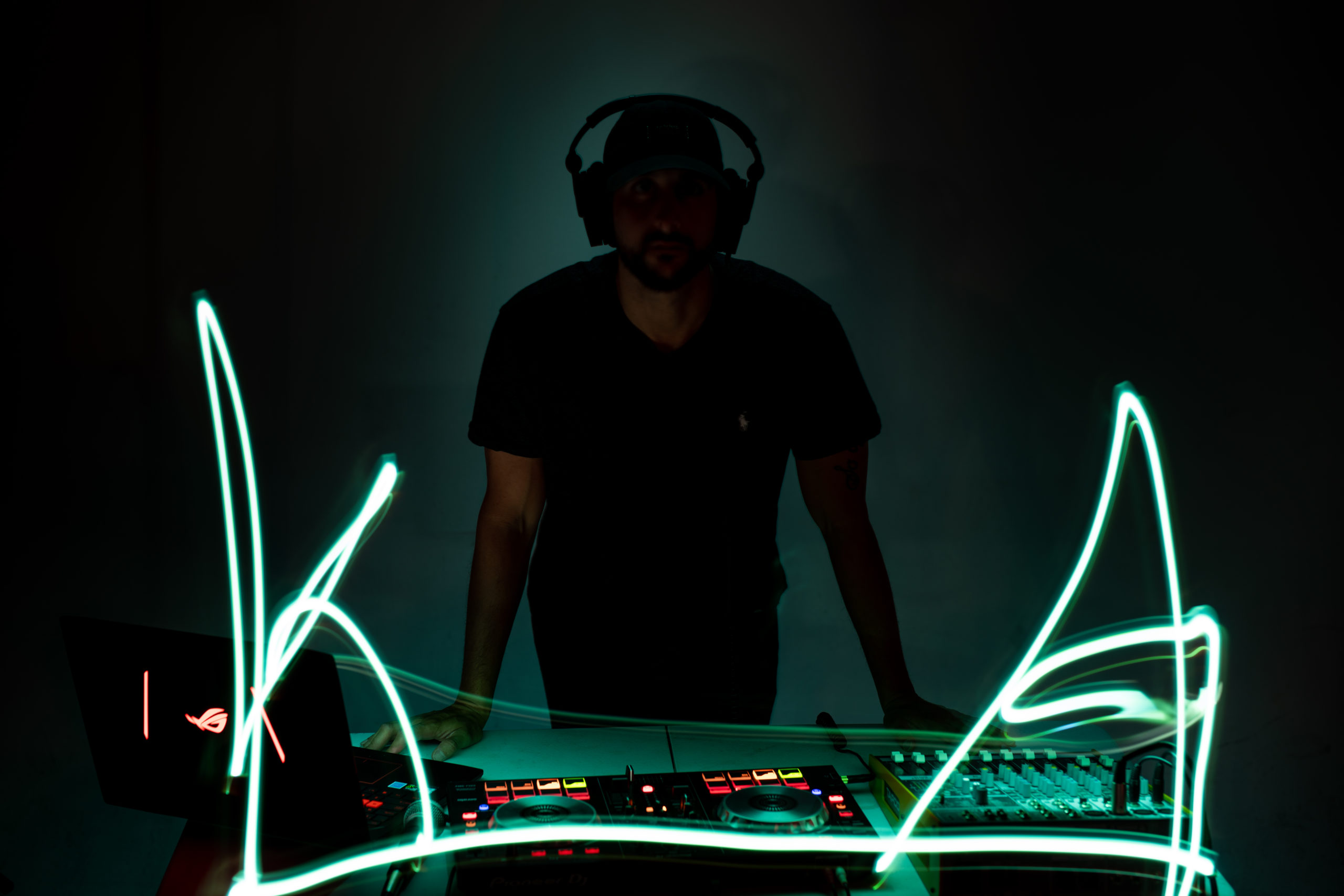 Man standing with black hat, beard and clothes looking towards camera behind a DJ machine with green neon lights. He is wearing headphones.
