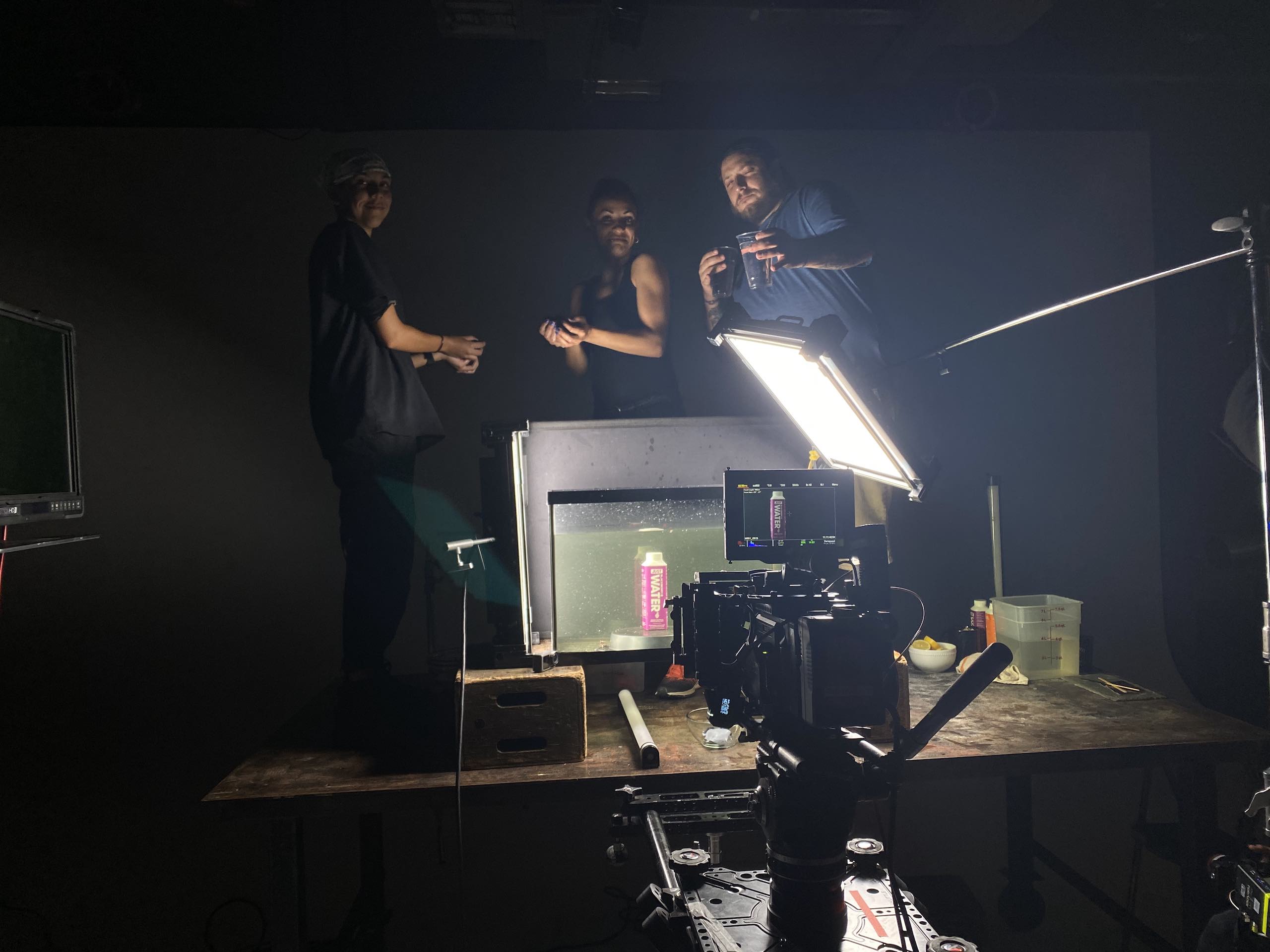 View from behind of camera trained on purple drink container in a tank filled with water in a studio with crew members standing by