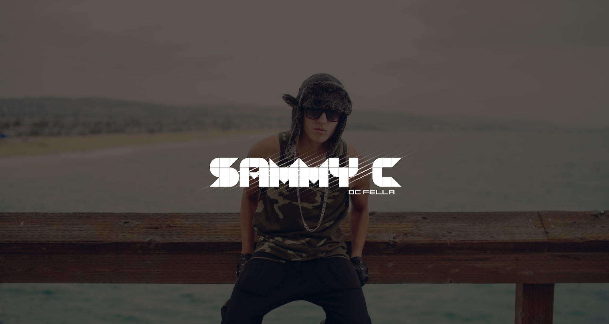 SammyC Artist Management Services White SammyC DC Fella logo on dimmed background of young man wearing a winter hat and camouflage tank top with chain, leather gloves and black pants