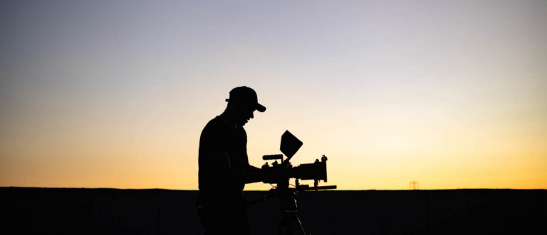 Silhouette side profile of crew member wearing cap using video camera at dusk