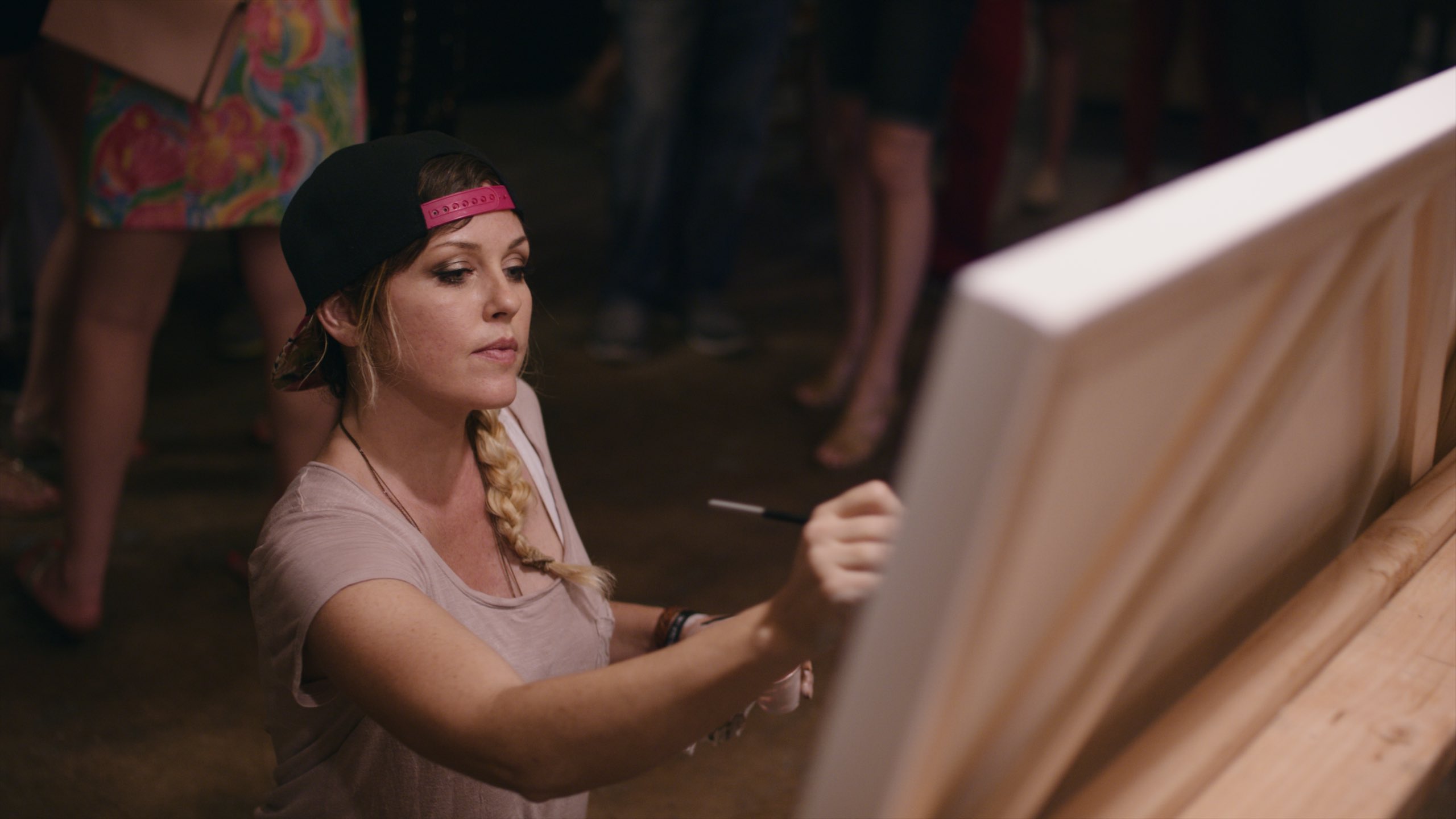 Art Battle International Woman with long blond hair wearing a black and pink cap on backwards painting on canvas with people behind her