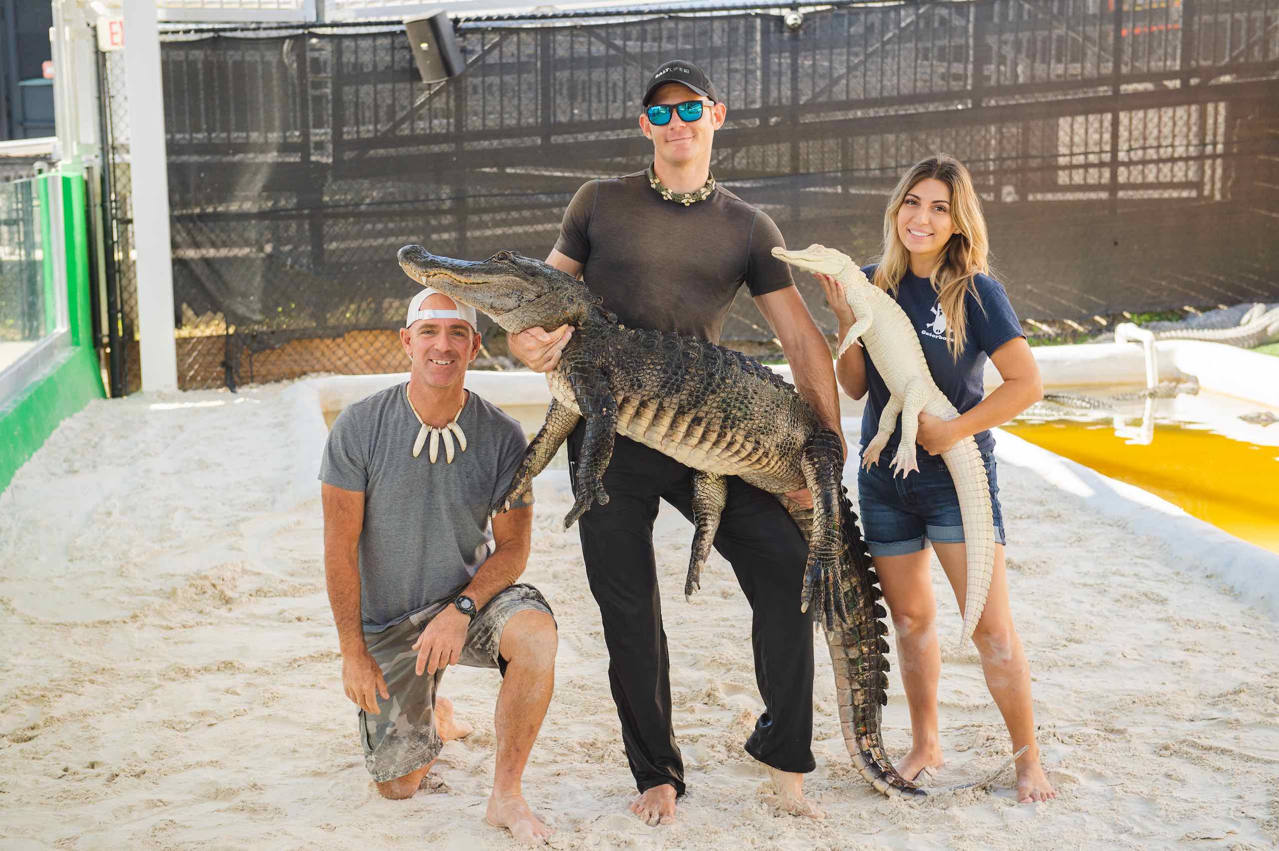 Everglades Holiday Park Two men with one carrying an alligator with a woman carrying a small albino alligator posing in the sand pit