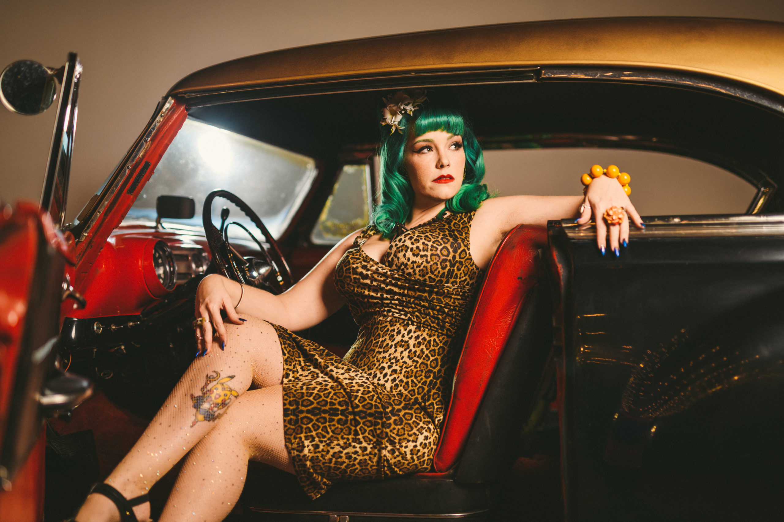 Closeup of woman wearing a green wig and leopard patterned dress and speckled pantyhose posing in a black car with red and black seats and trim looking off to the side
