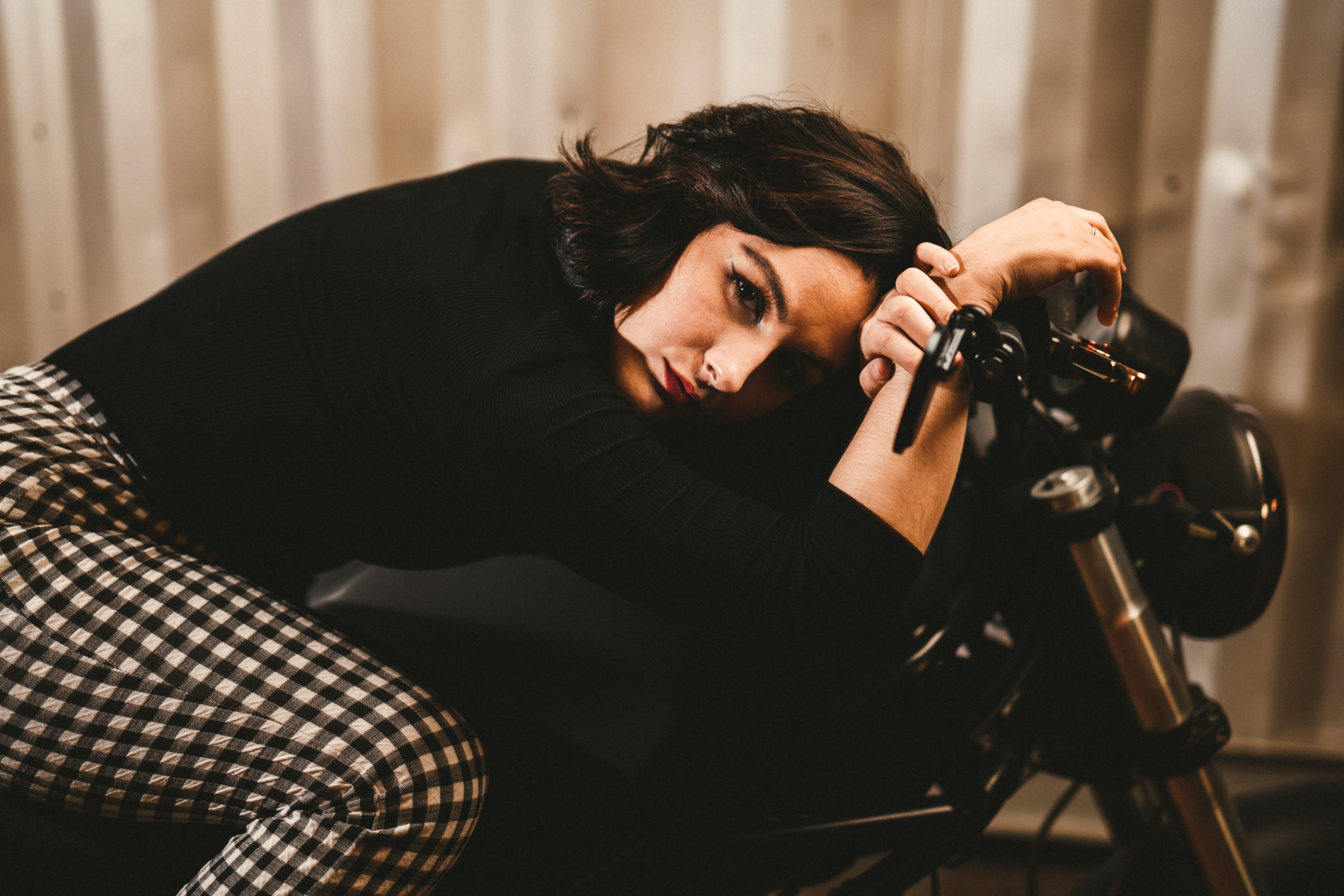 Closeup of woman with short brown hair wearing black top and black and white checkered pants posing for the camera crouching down on a motorcycle