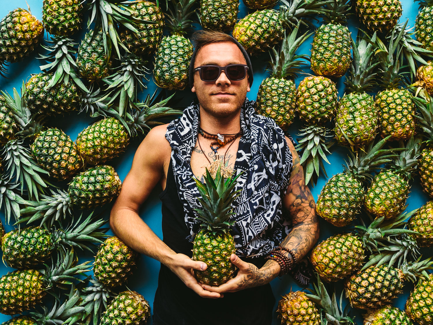 Artwalk 2018 May Pineapple Pyramid Man wearing a black knit cap smiling posing for camera holding a pineapple with pineapples surrounding his head on a teal background