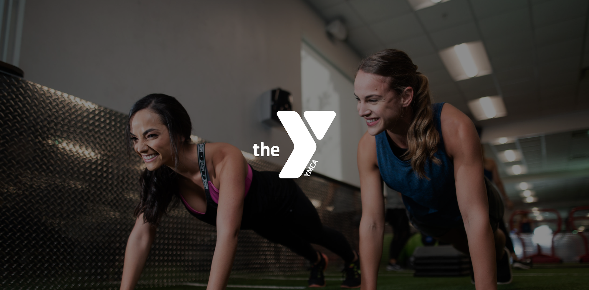 White The Y logo with background of two young women with long hair doing pushups at the YMCA.