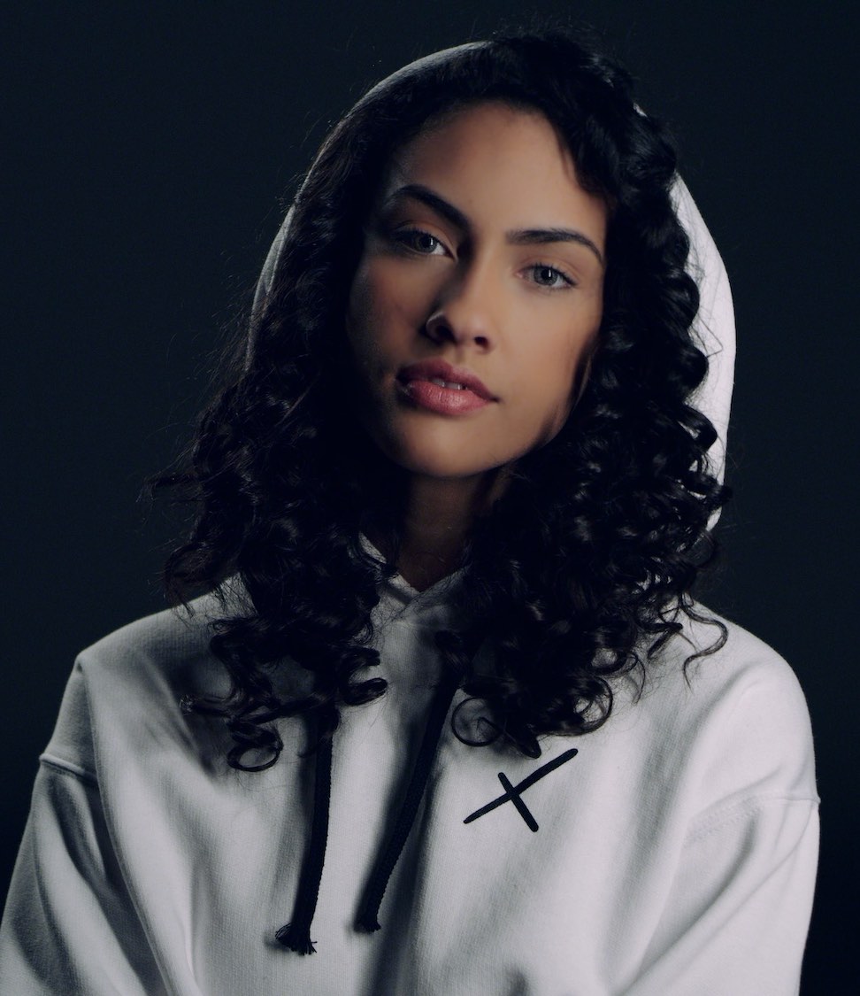Headshot of female model with curly black hair posing for camera wearing a white sweatshirt with black Uncreative logo with hoodie over her head