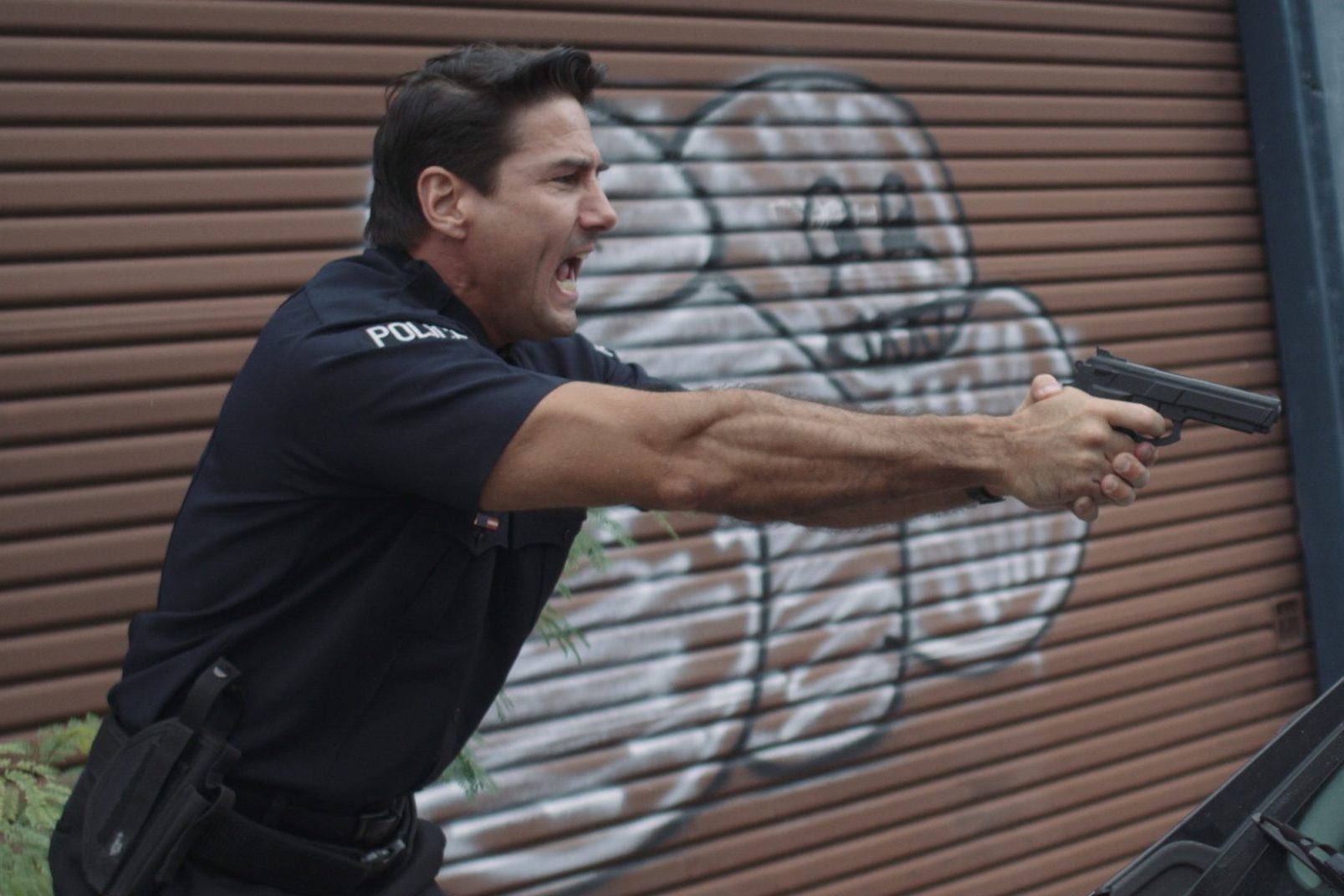 Side profile of police officer wearing uniform holding a gun in outstretched hands shouting with graffiti on wall behind him