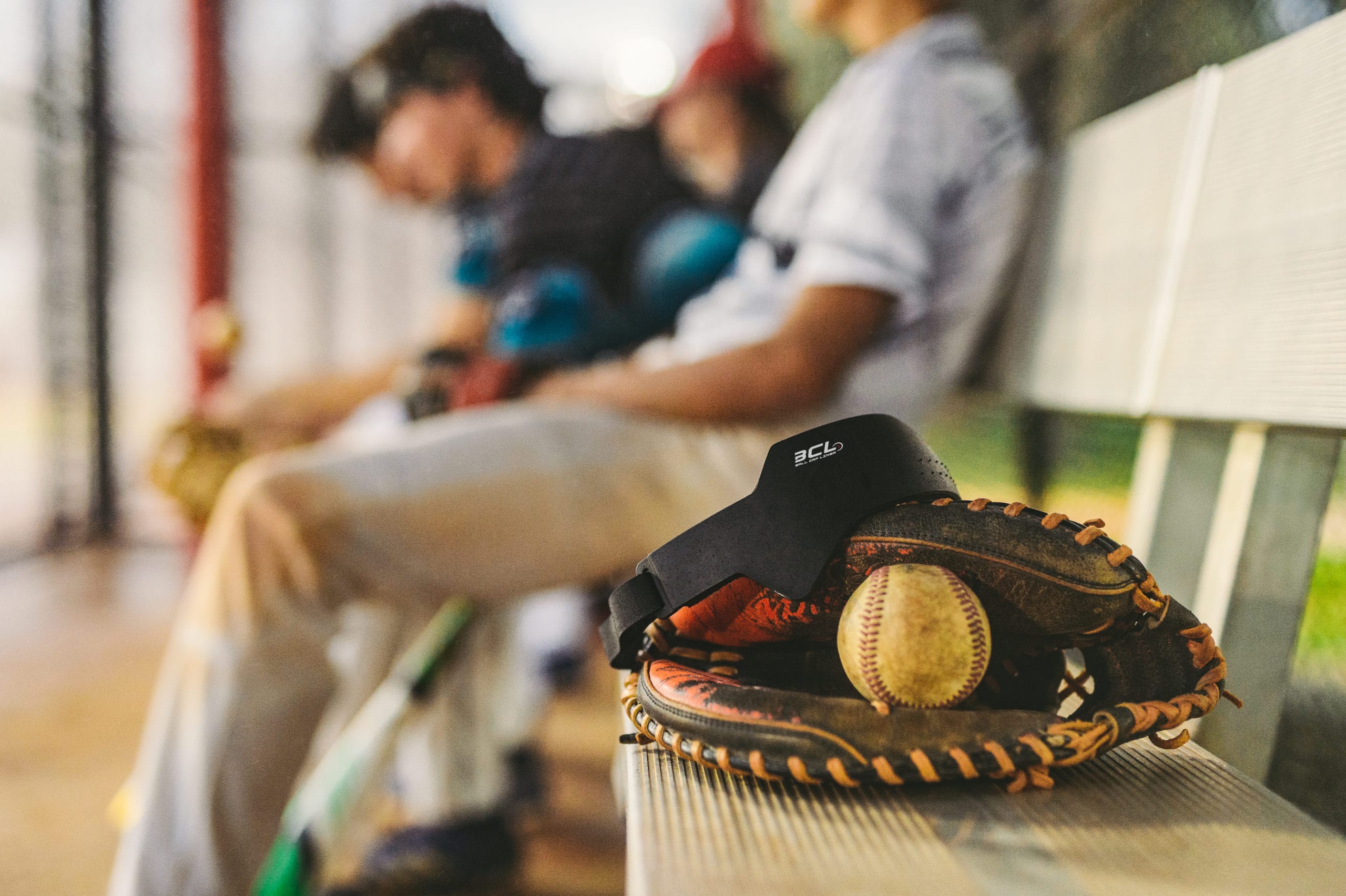 Closeup of baseball mitt with baseball in it on a bench with Ball Cap Liner on it with two male baseball players sitting nearby