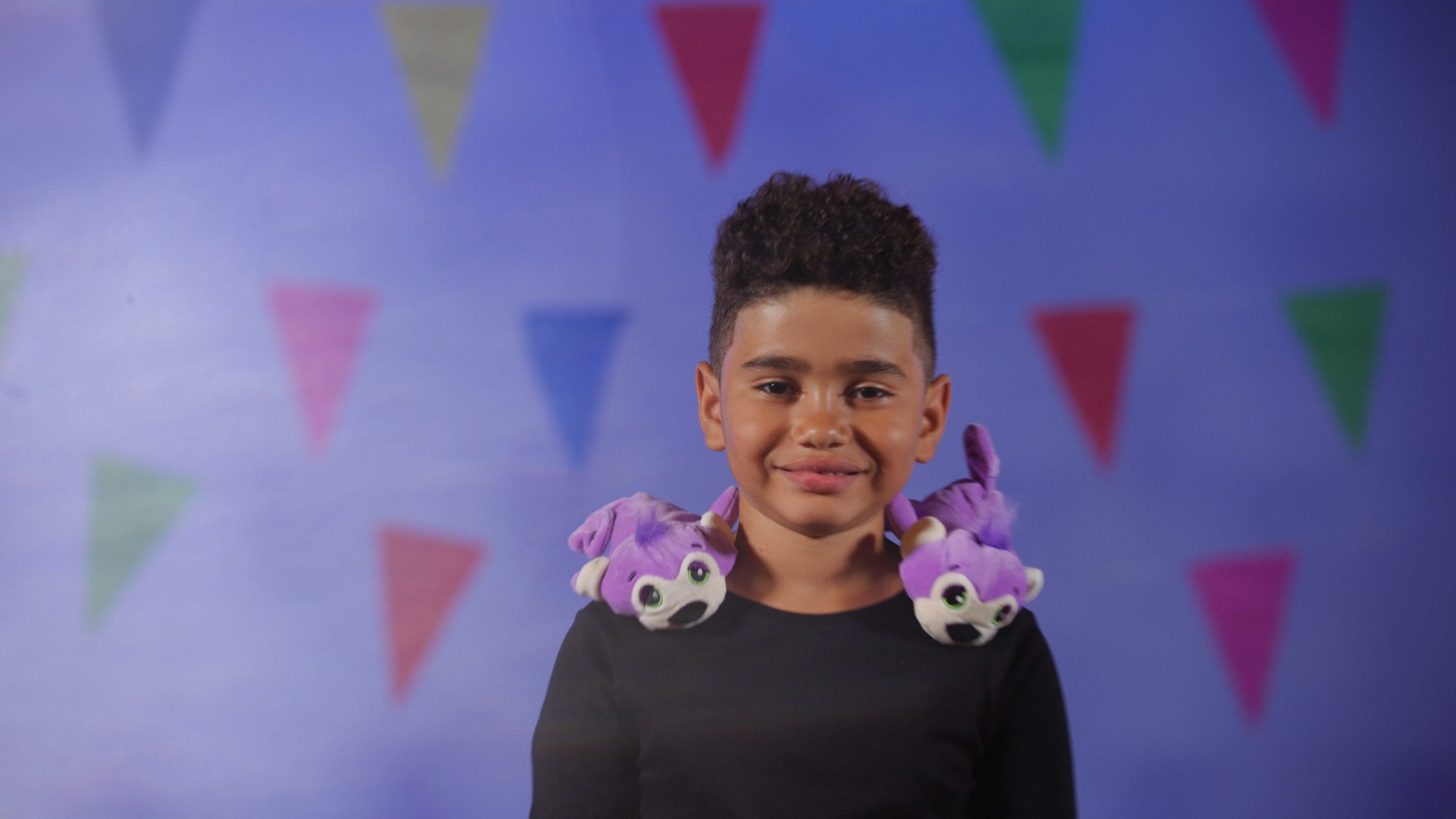 Male child posing for camera smiling with two purple stuffed animals on his shoulders