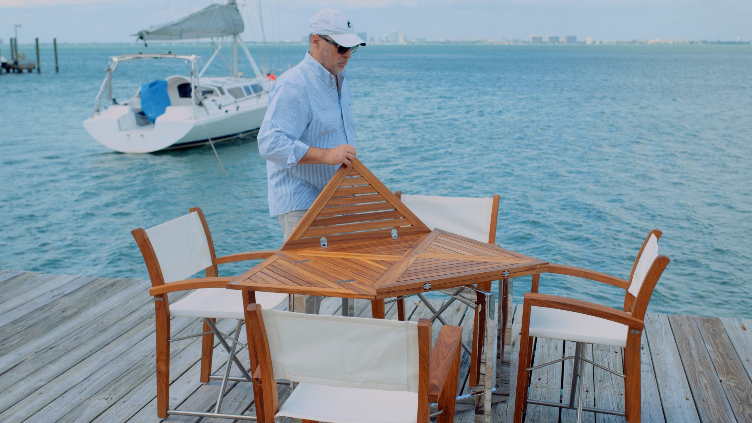 Man wearing white cap and light blue shirt setting up table and chairs on a pier with a boat in the background on the water
