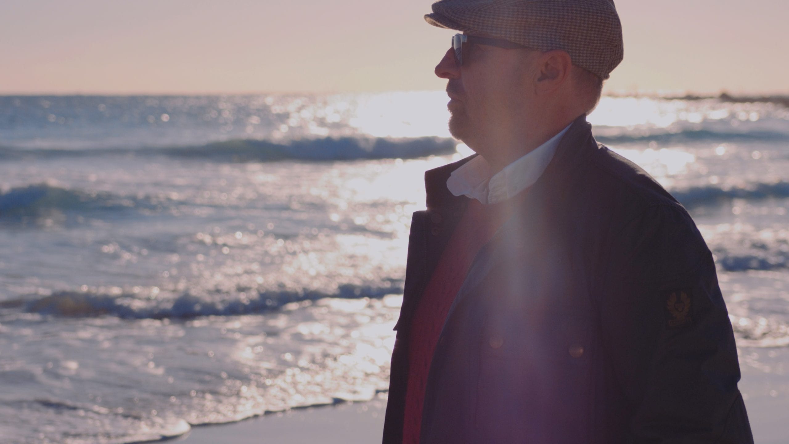 Side profile of man wearing a cap, sunglasses, red sweater and blue jacket standing on a beach