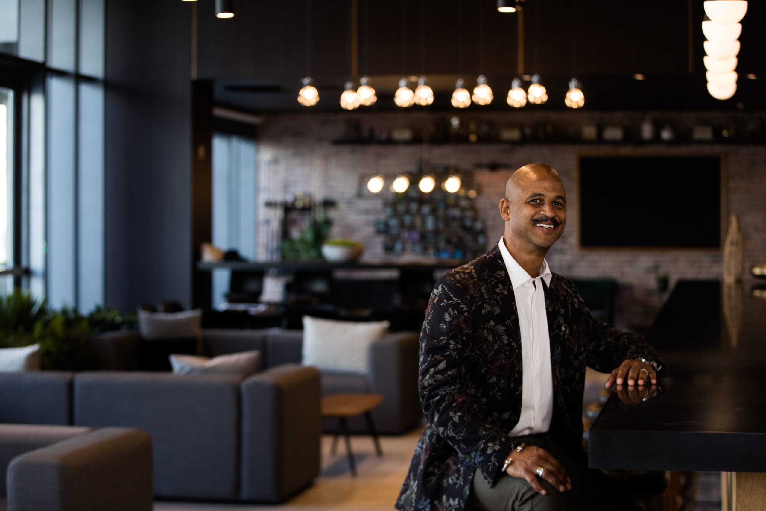 Fort Lauderdale Illustrated Men of Style Bald African American male model with moustache wearing gray jeans, white shirt and multicolored jacket sitting by a bar smiling