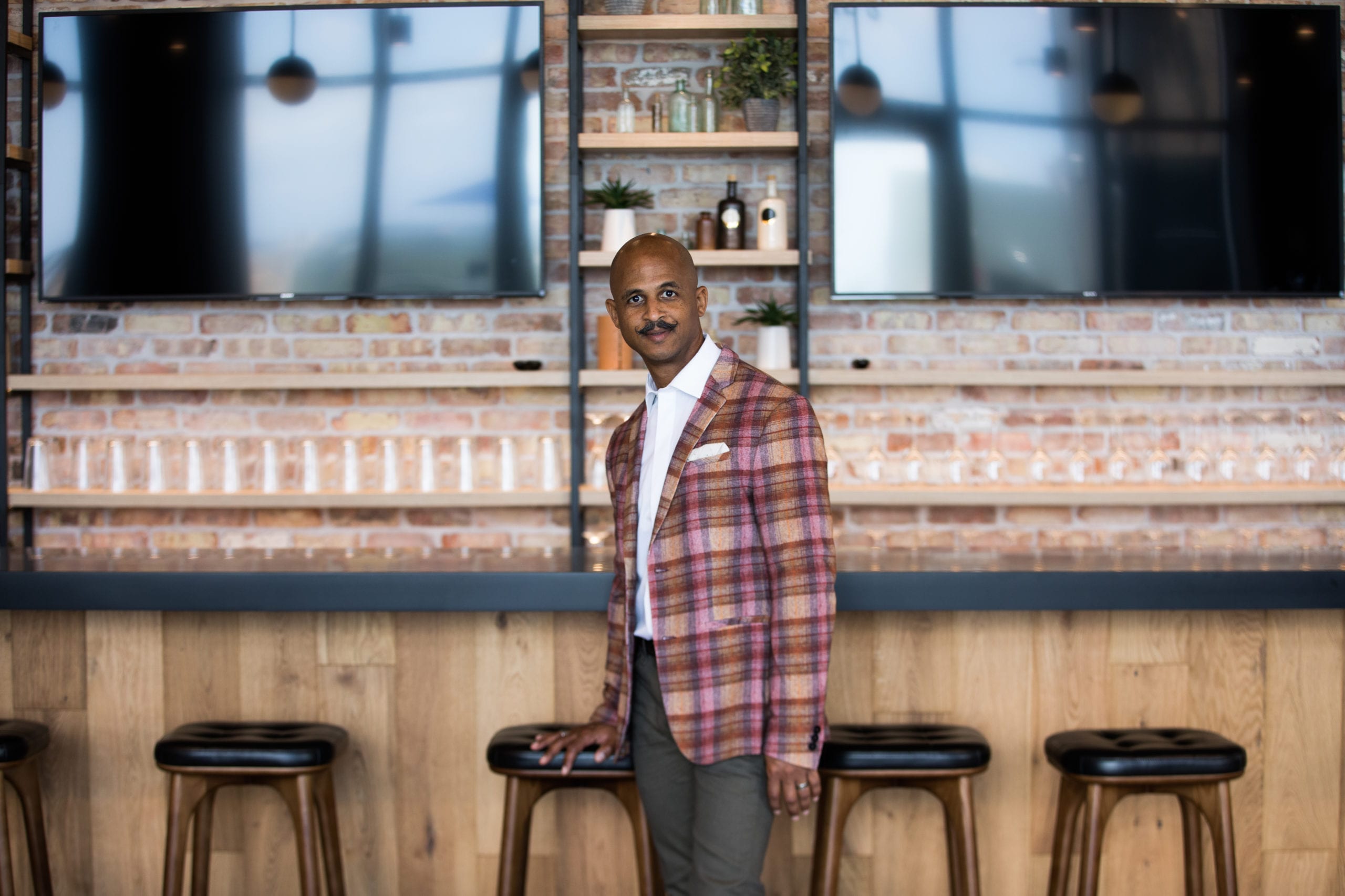 Fort Lauderdale Illustrated Men of Style Bald African American male model with moustache wearing gray jeans, white shirt and multicolored jacket leaning up against a bar