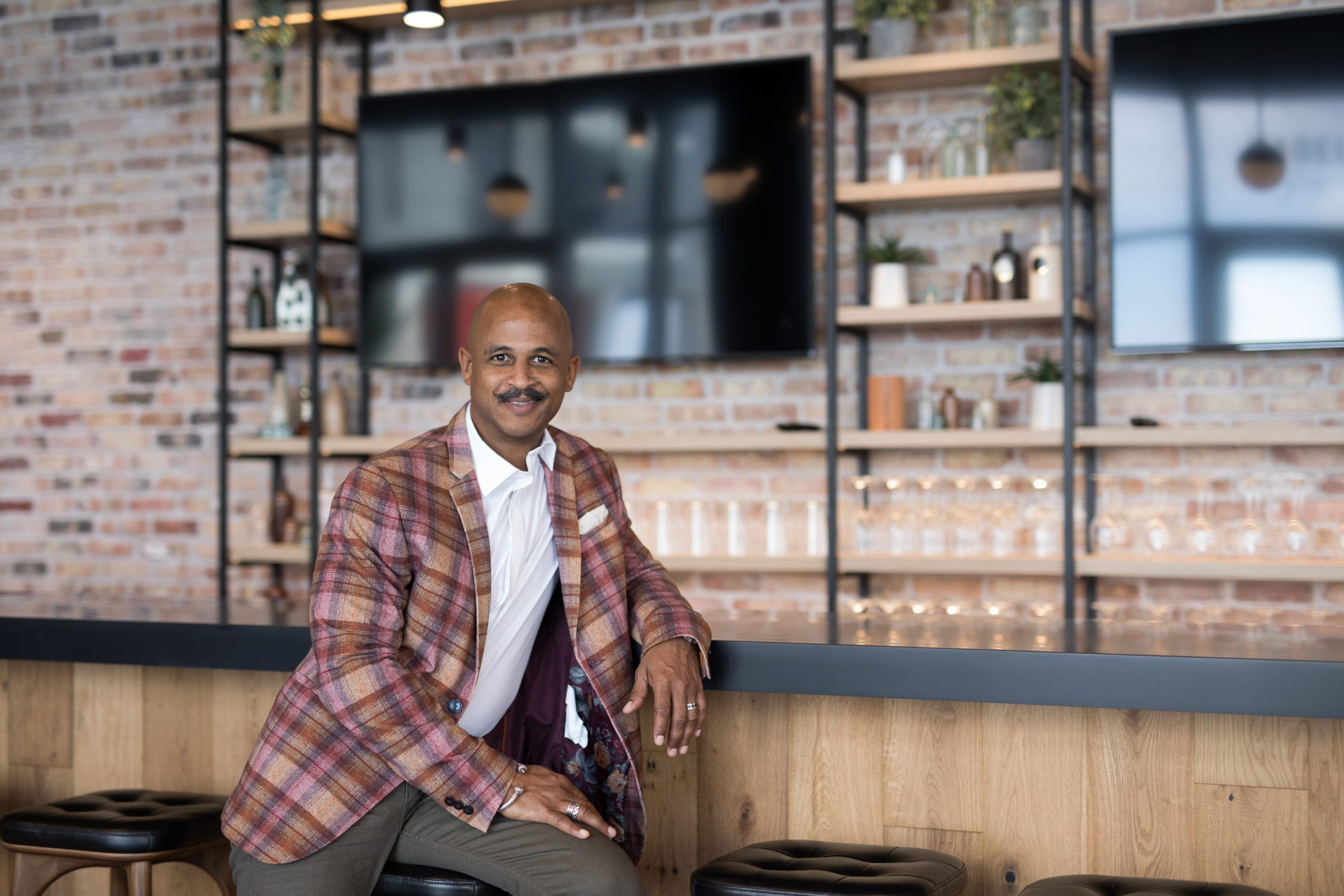 Fort Lauderdale Illustrated Men of Style Bald African American male model with moustache wearing gray jeans, white shirt and multicolored jacket sitting up against a bar smiling