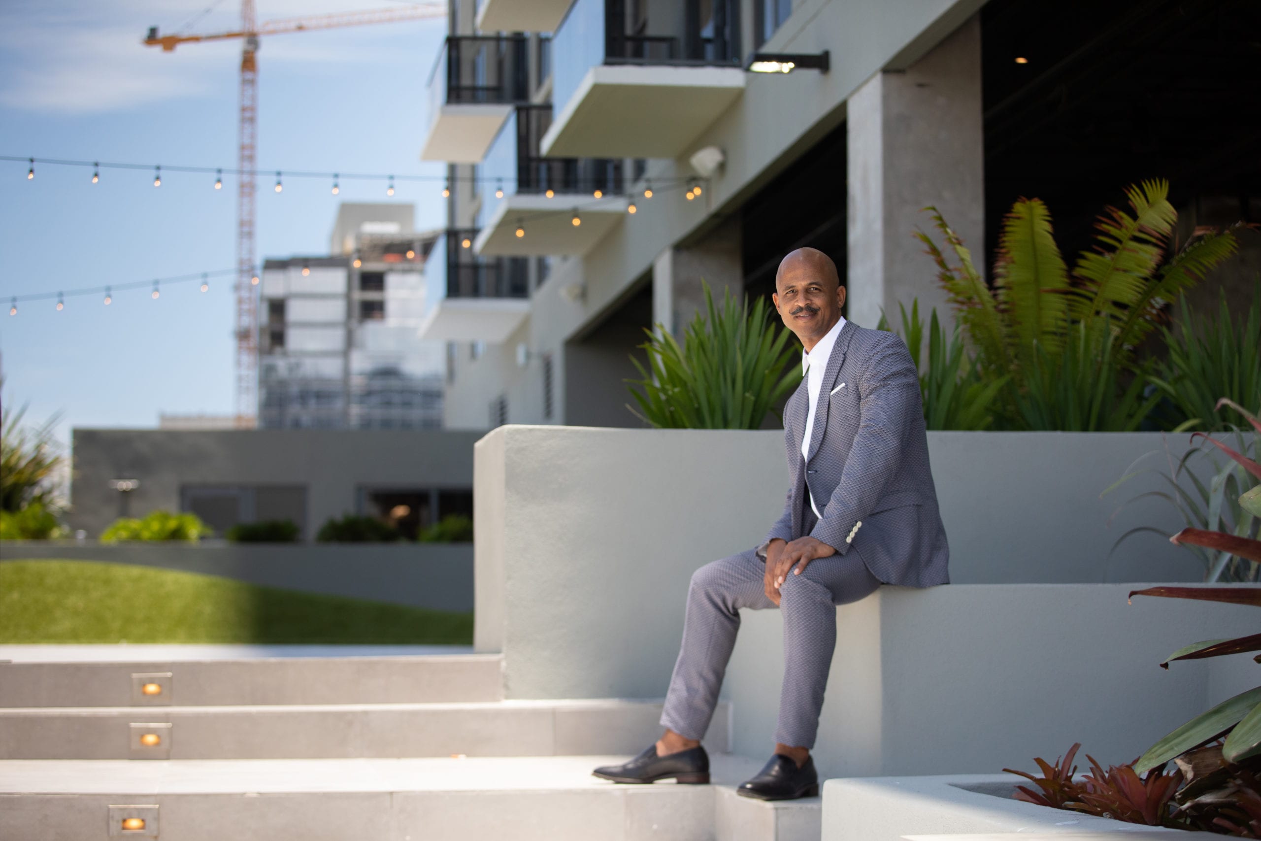 Fort Lauderdale Illustrated Men of Style Bald African American male model with moustache wearing gray suit and white shirt sitting near stairs