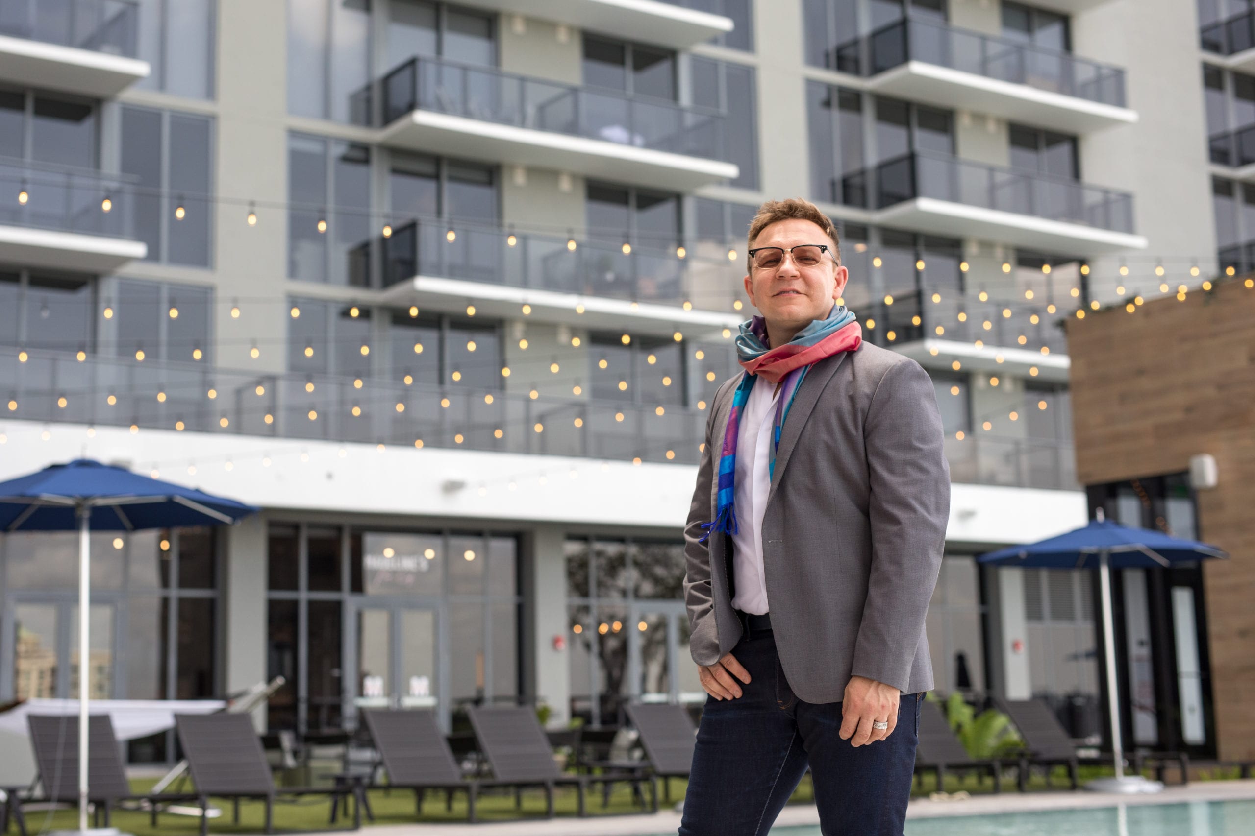Fort Lauderdale Illustrated Men of Style Side profile closeup of man wearing glasses, gray suit jacket and jeans and colorful scarf posing for the camera standing in front of a building
