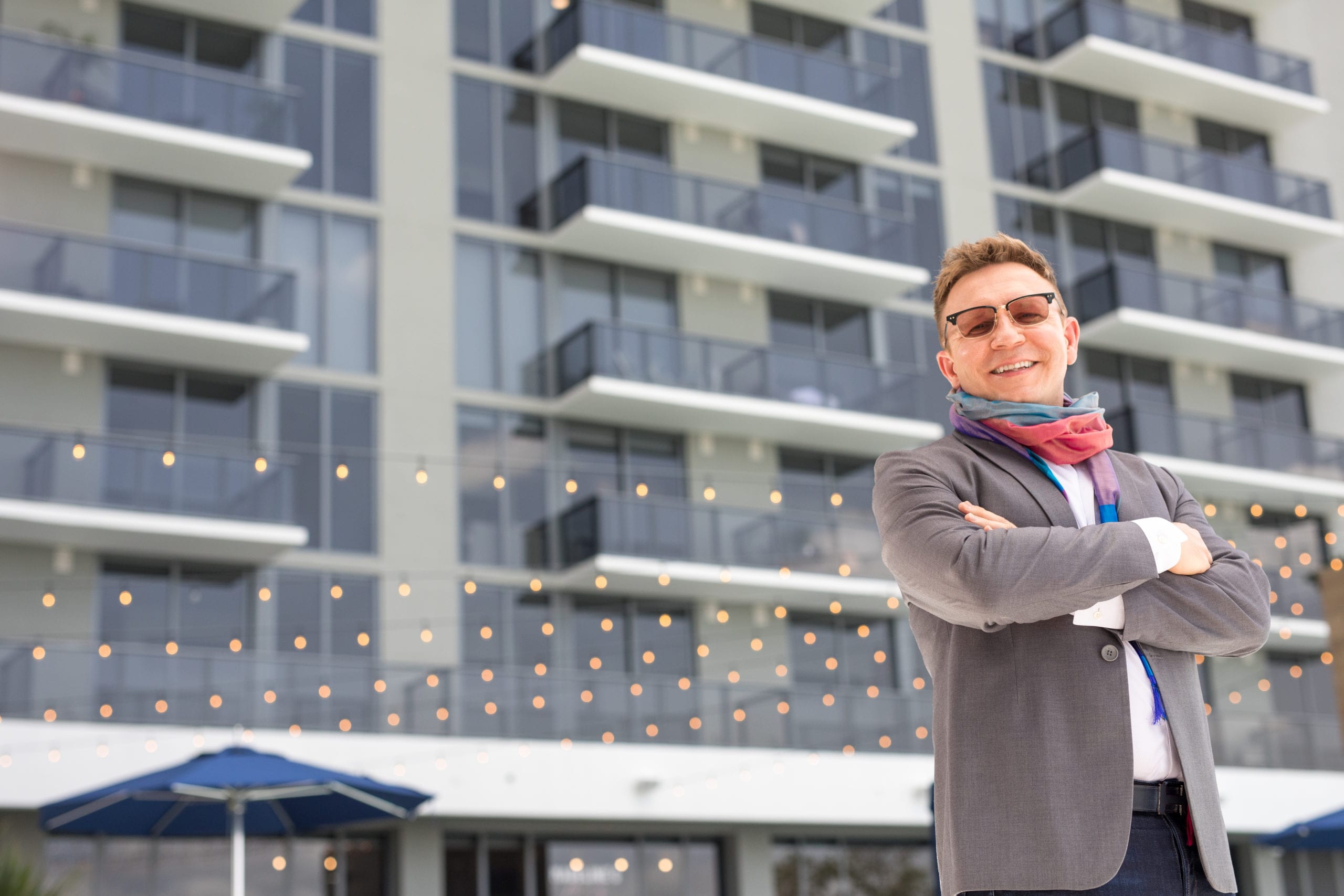 Fort Lauderdale Illustrated Men of Style Closeup of man wearing glasses, gray suit jacket and jeans and colorful scarf posing for the camera standing in front of a building