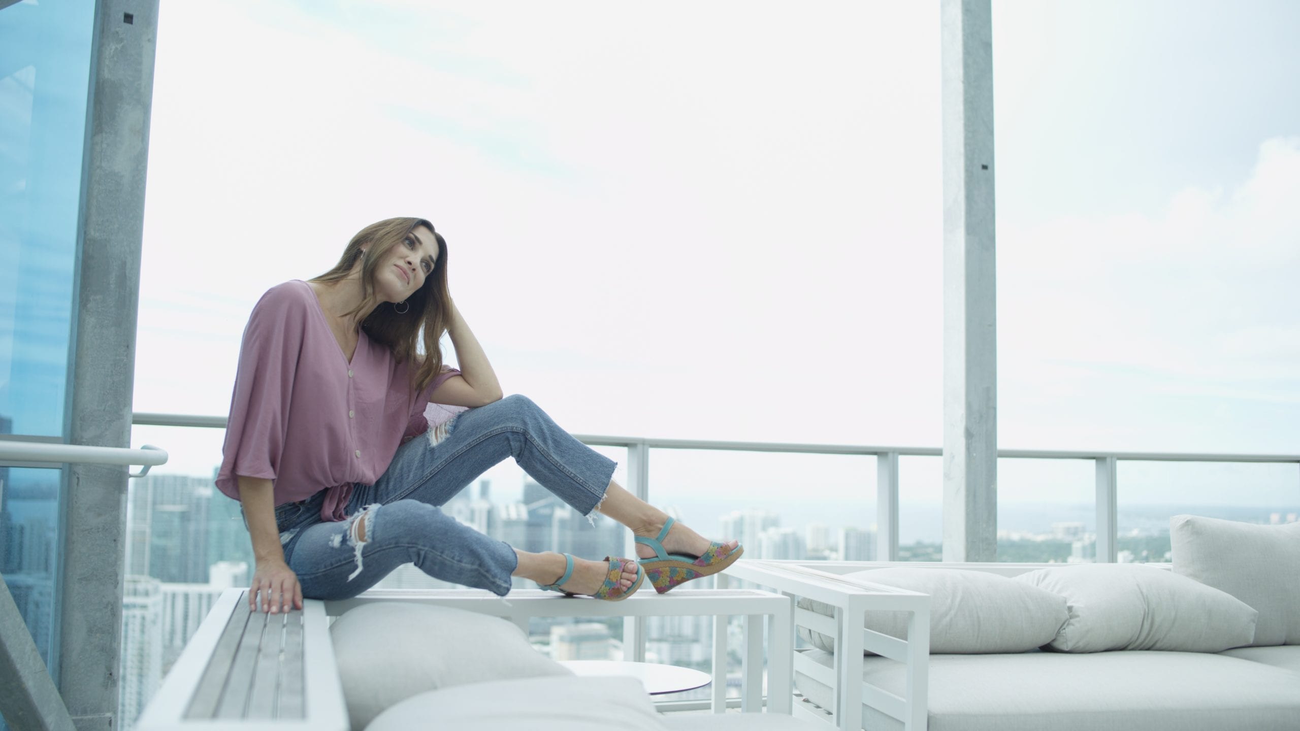 Spring Step Side profile of Lartiste Pranav posing for camera wearing light purple top and jeans with heels sitting on edge of patio furniture