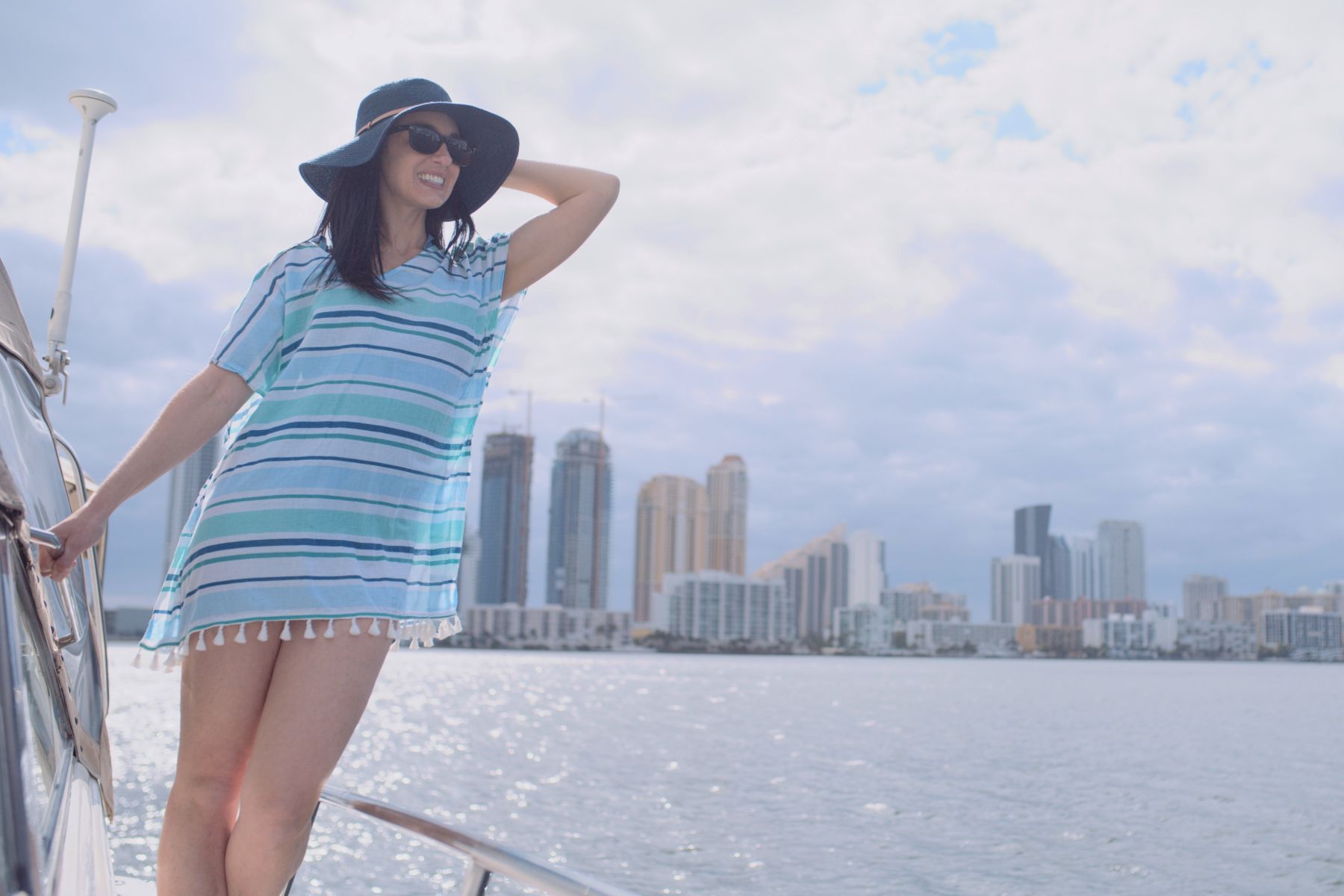 Macken Koya Bay Woman wearing hat, sunglasses and a multicolored blue top posing on a yacht with the city coastline in the background
