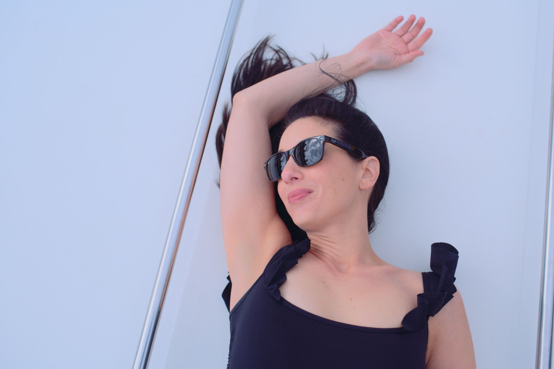 Macken Koya Bay View from above of woman wearing a black bathing suit and sunglasses sunbathing on a white yacht with her arm over her head