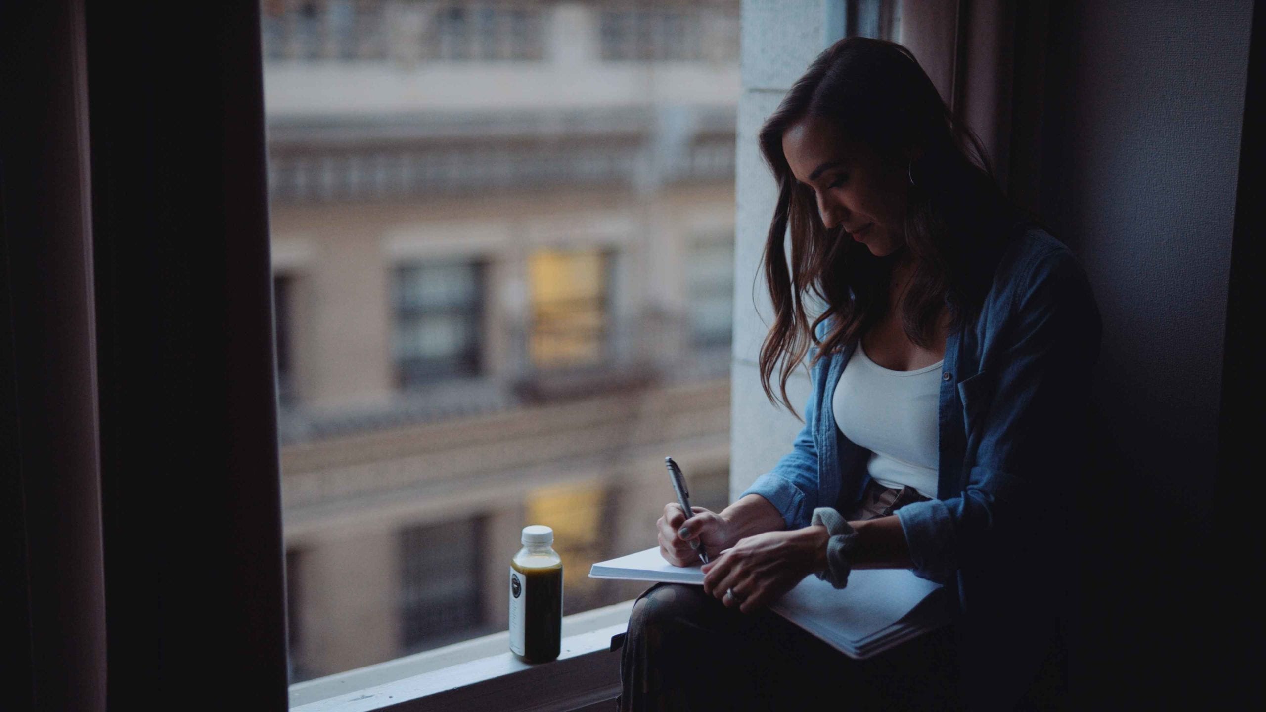 Woman with long hair sitting on a window ledge writing into a notebook