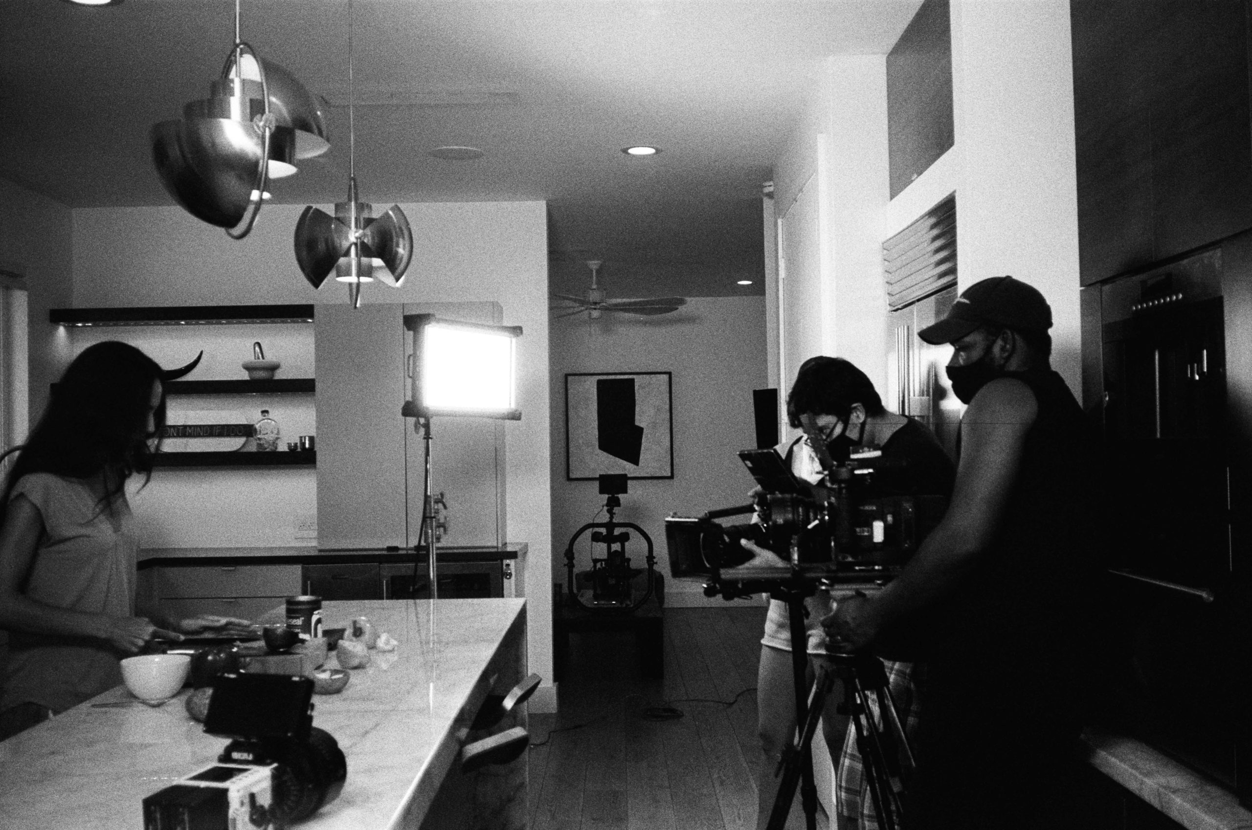 Black and white photo of a cooking show in progress with a woman cooking at the counter and two males wearing masks filming the show. There is another video camera on the counter as well.
