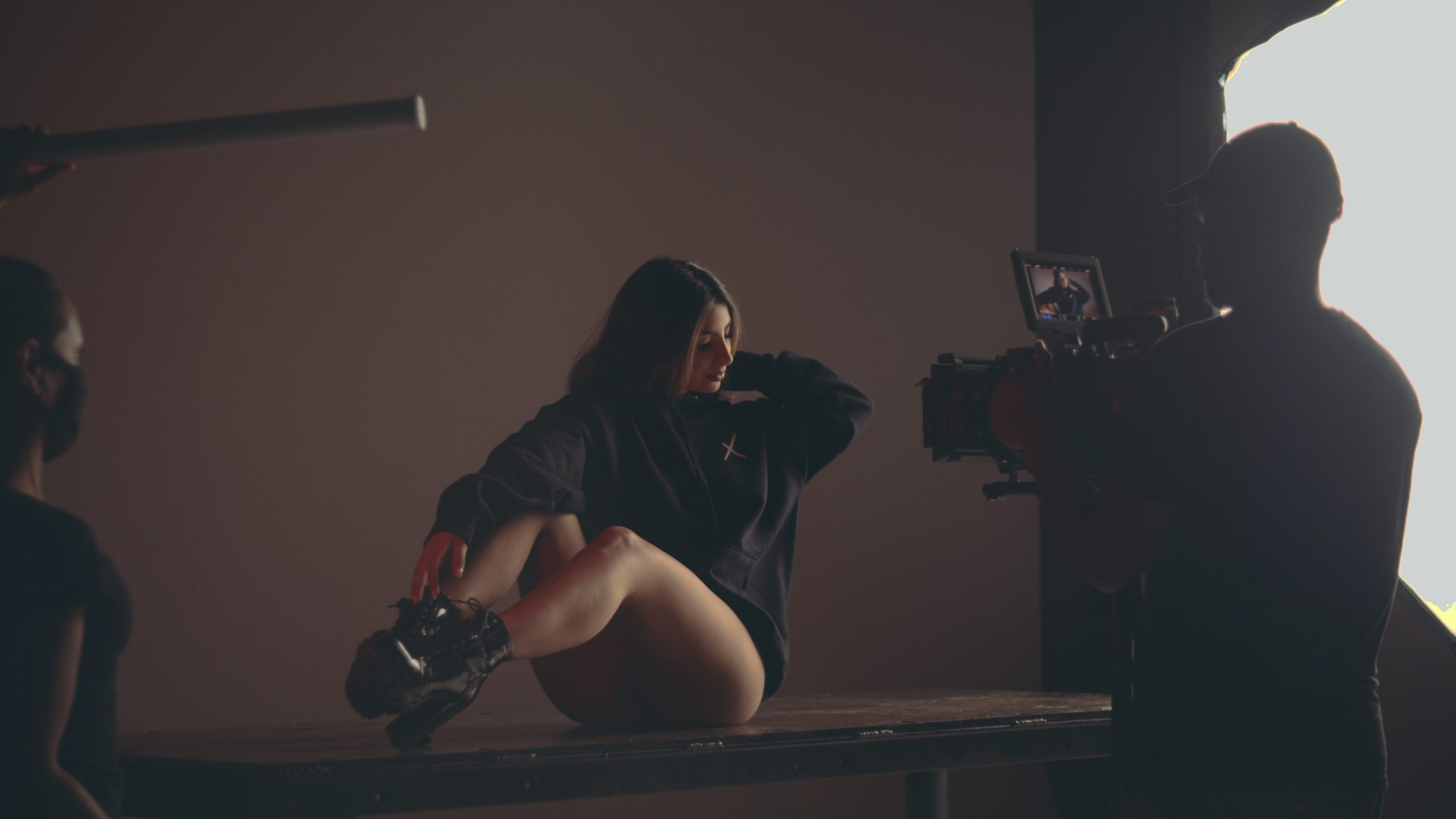 Uncreative Shop Anna posing for the video camera sitting on a table wearing a black pullover, thong and shoes with two crew members looking on