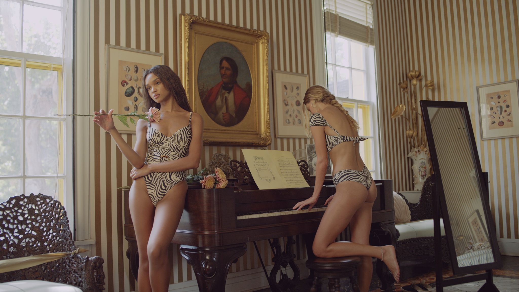 Two female models wearing black and white swim suits in a room with one holding a flower leaning against the piano and the other playing the piano