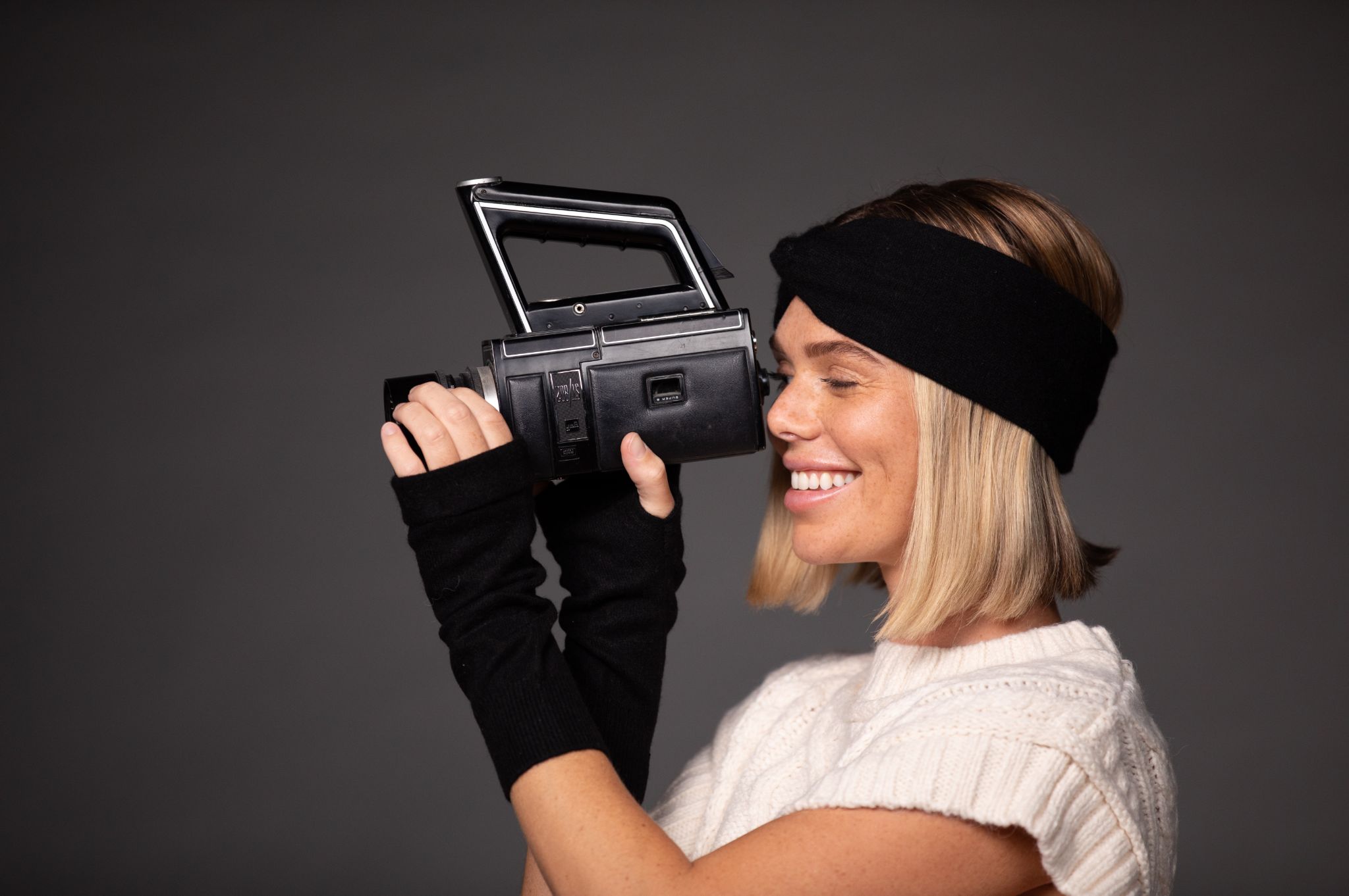 Side profile of woman with long blond hair smiling and using a video camera wearing a black headband, hand sleeves and white cropped sweater against a gray backdrop
