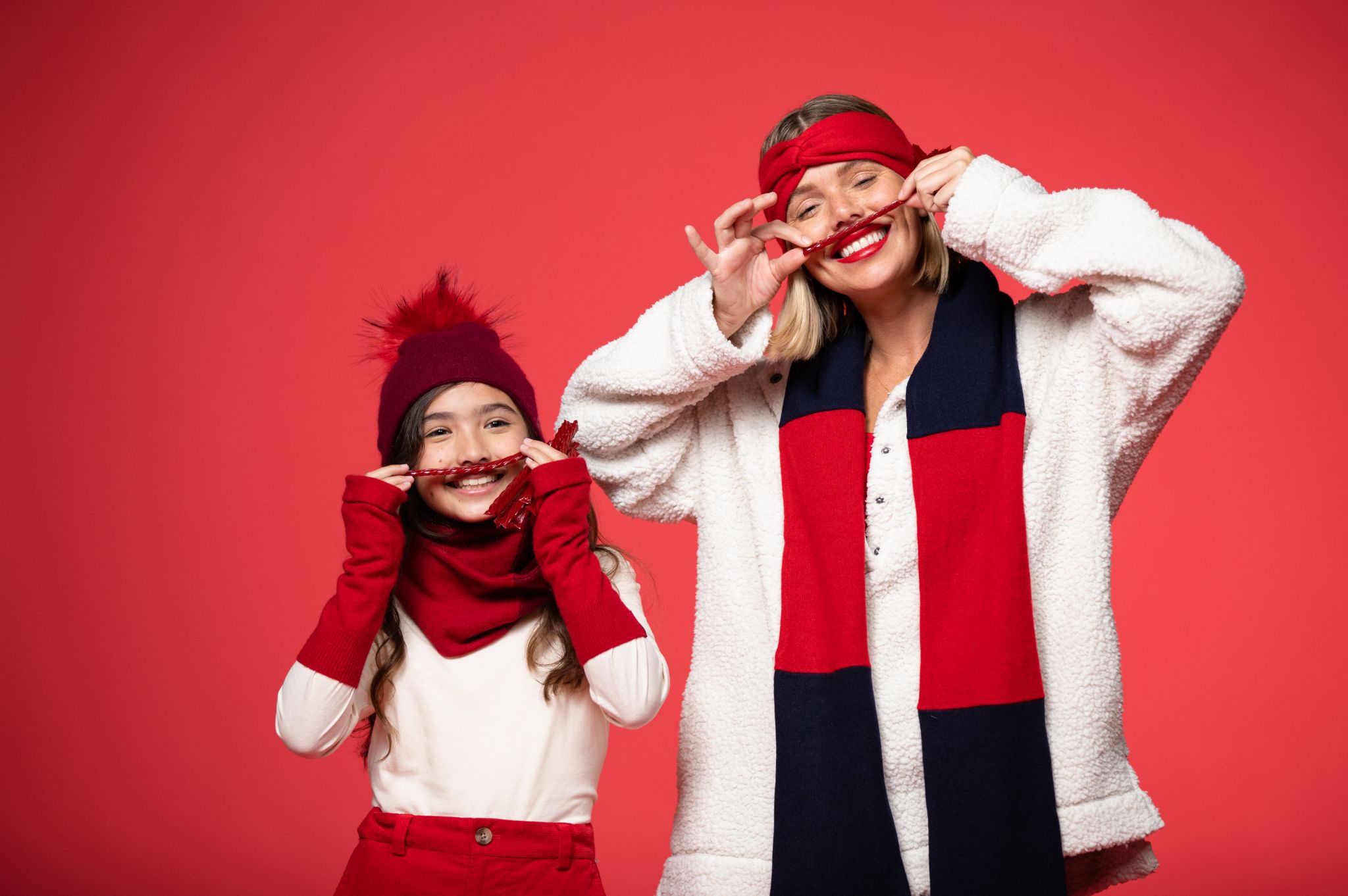 Girl model wearing red headband, scarf and pants along with white top and female model wearing red hand band, white jacket and black and red scarf against a red backdrop