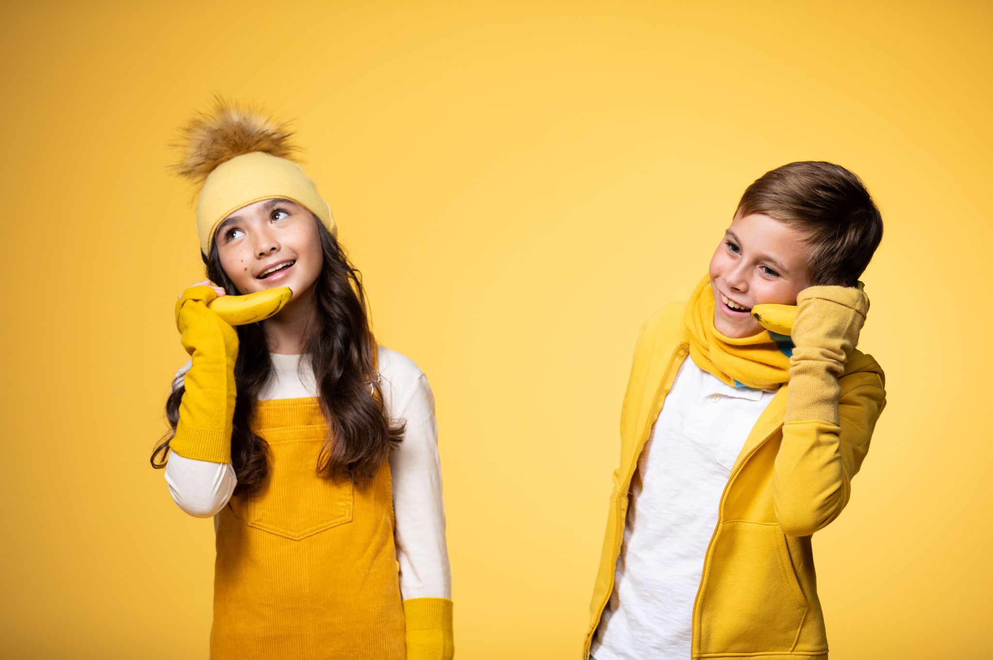 Boy model wearing a yellow scarf. jacket and white top and girl model wearing a yellow headband, hand sleeves and overalls as well as white top posing for the camera both using bananas as phones against a yellow backdrop