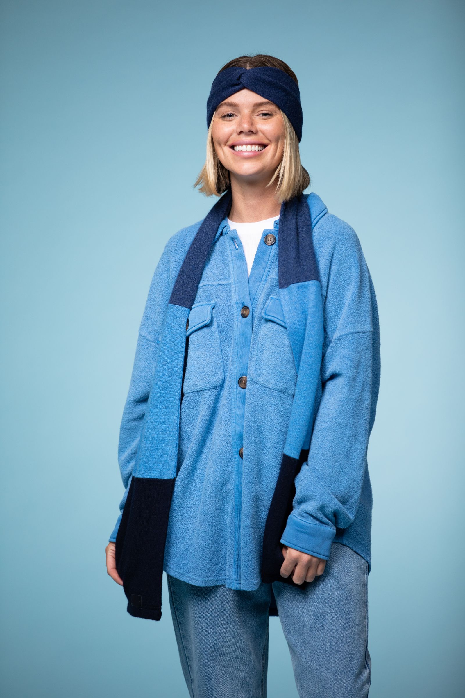 Female model wearing a dark blue headband, dark blue and blue scarf, light blue sweater and white top as well as jeans smiling and posing for the camera against a light blue backdrop