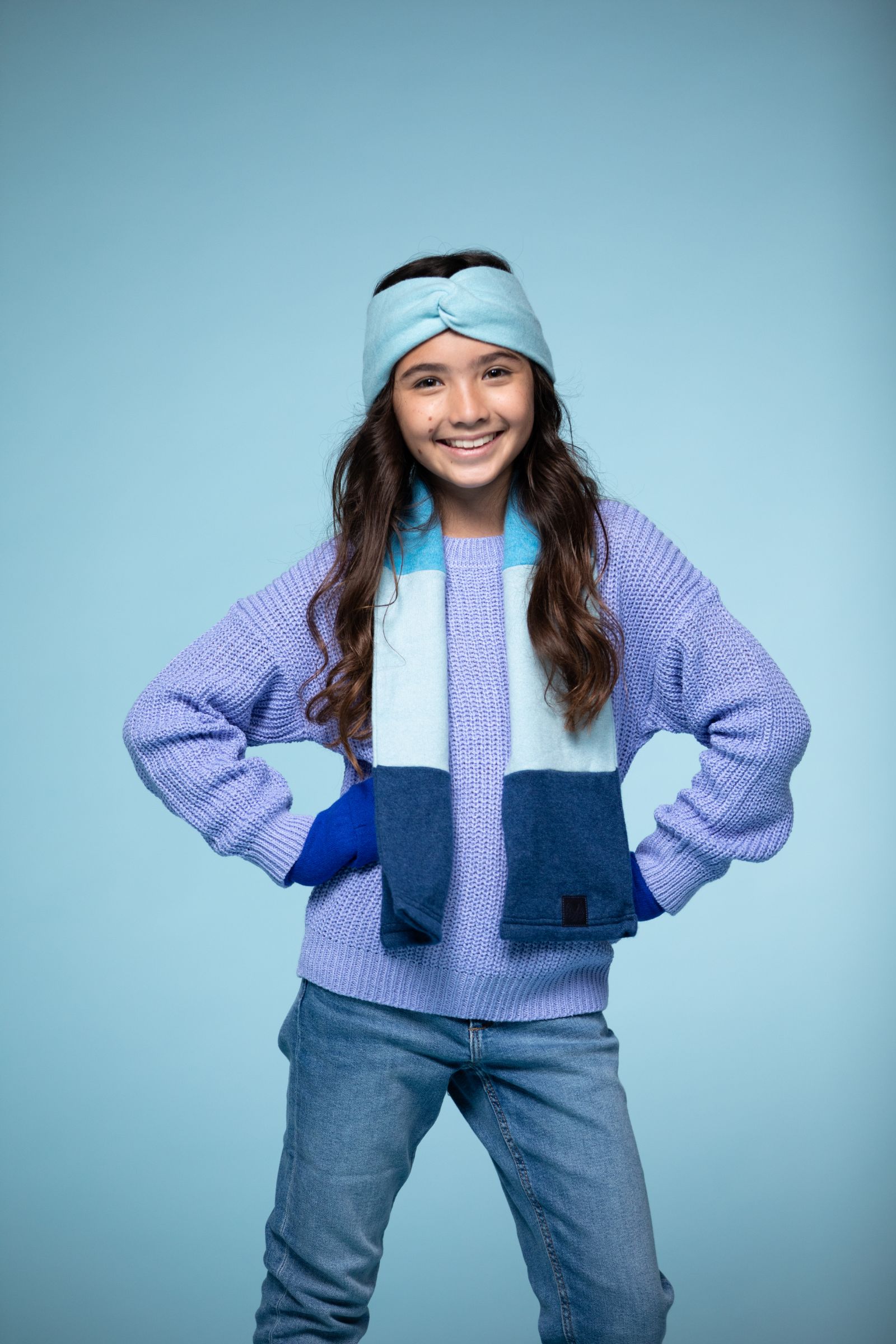 Girl model wearing a light blue head band, multicolored scarf, purple sweater and jeans smiling and posing for the camera against a light blue backdrop