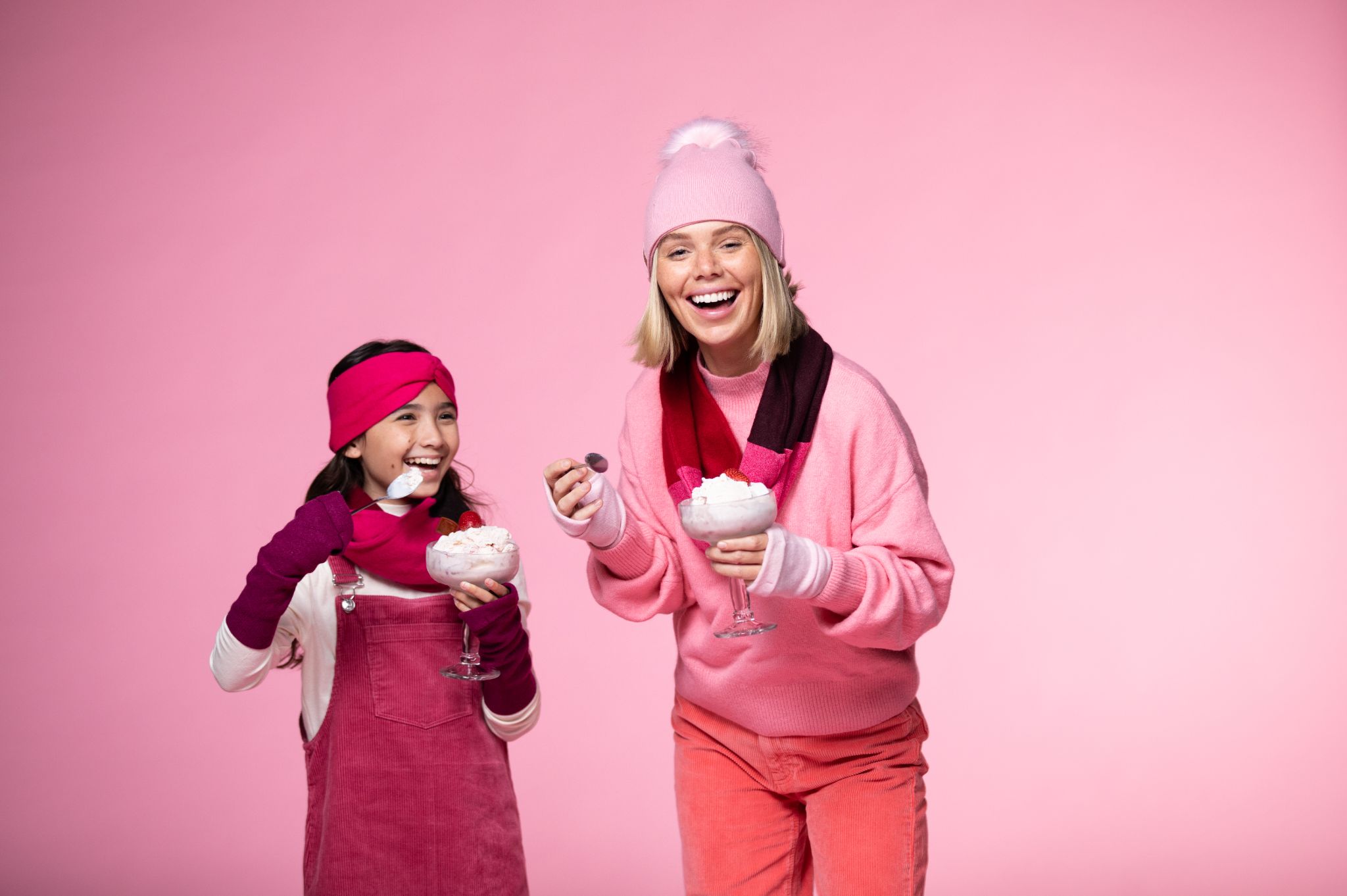 Girl and female models posing for the camera smiling holding ice cream sundaes wearing red and pink outfits respectively against a pink backdrop