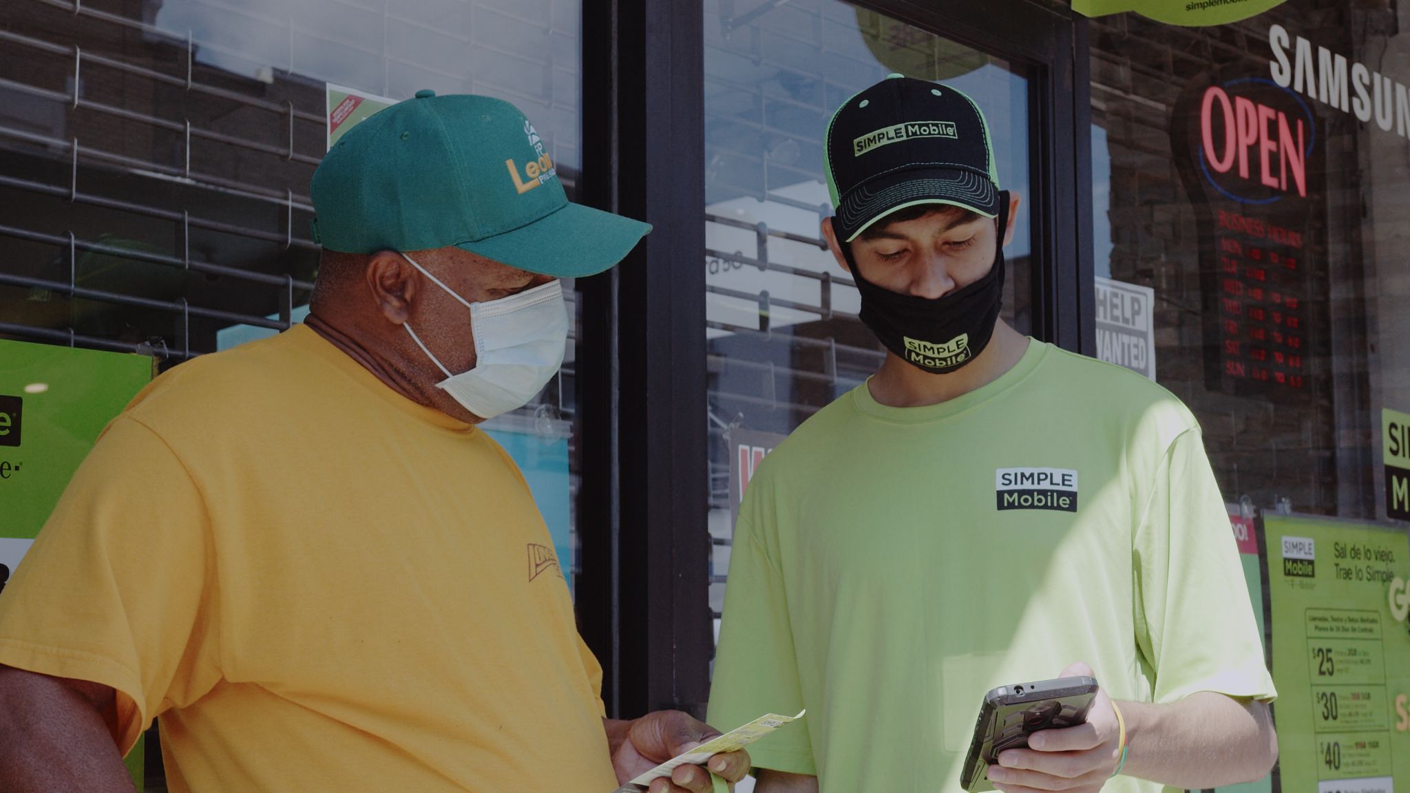 Male Simple Mobile employee wearing black mask and cap showing something on a call phone to a customer wearing a yellow shirt, surgical mask and cap