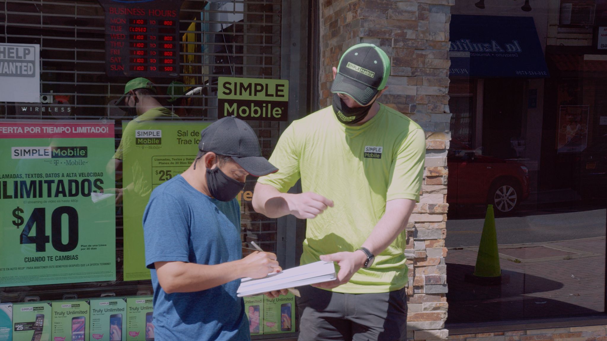 Simple Mobile employee wearing a black mask with logo having a paper signed by a customer wearing a black cap and blue shirt by the storefront