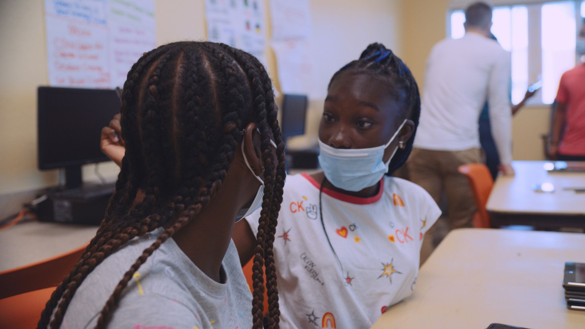 Two African American girls with braids and wearing surgeon masks facing each other in a classroom