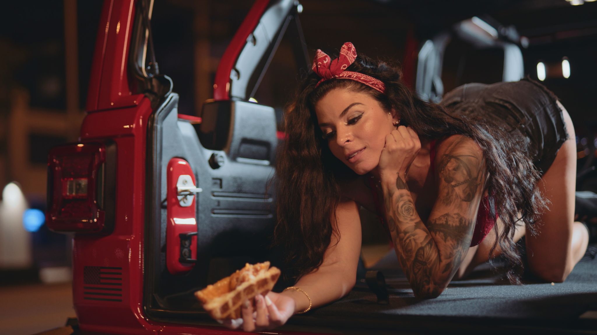 Woman with long brown hair wearing a red lacy bra with black shorts on her hands and knees in the back of a red jeep looking at a grilled cheese sandwich she is holding