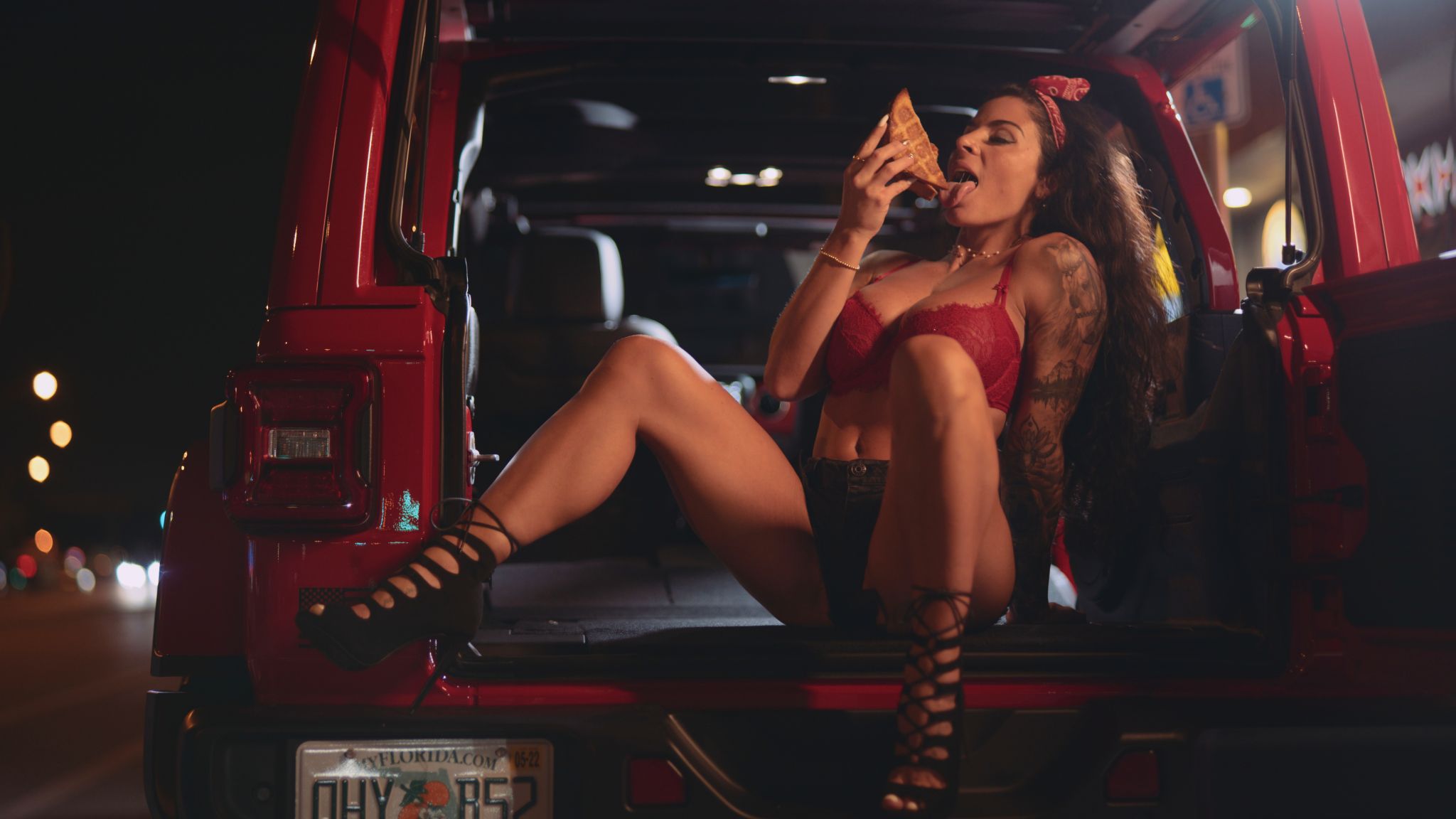 Woman with long brown hair wearing a red lacy bra with black shorts sitting in the back of a red jeep eating a grilled cheese sandwich