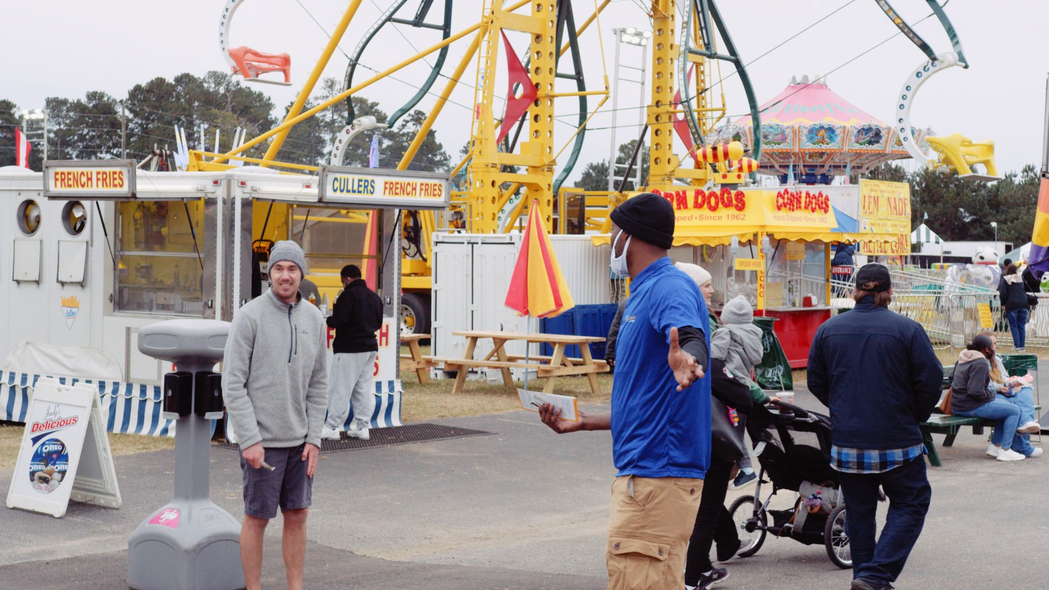 Man wearing a gray knit cap, sweatshirt and shorts smiling for the camera at a fair with other people nearby