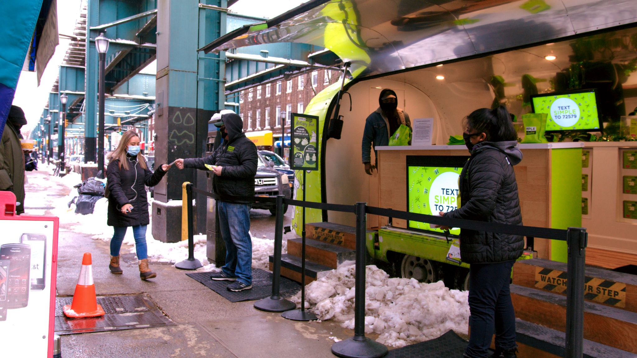 Storefront in a parked van with employees around and one giving a business card to female passerby wearing a surgical mask