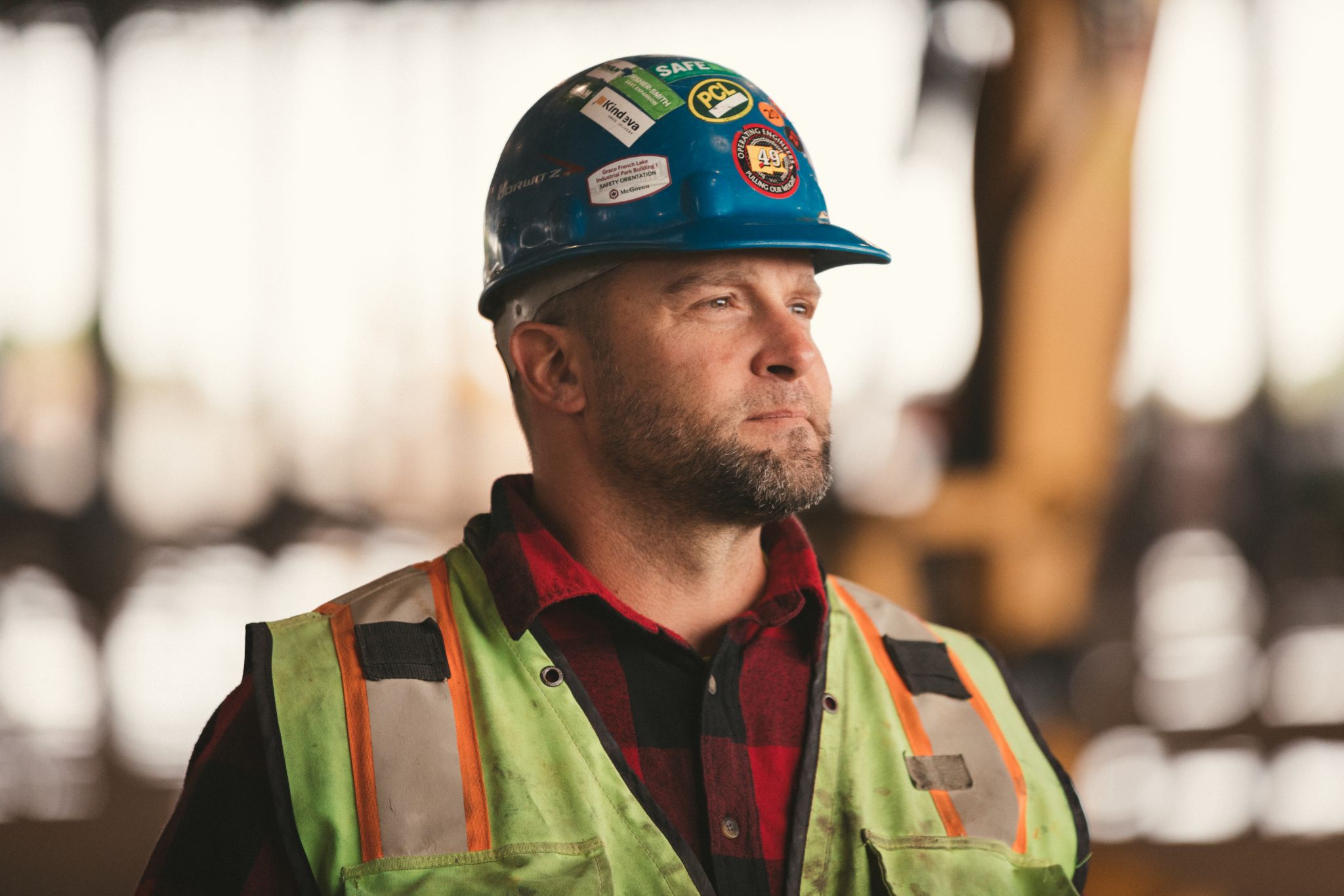 Closeup headshot of man with a short beard and hair wearing a red flannel shirt, safety vest and blue hardhat looking off to the side