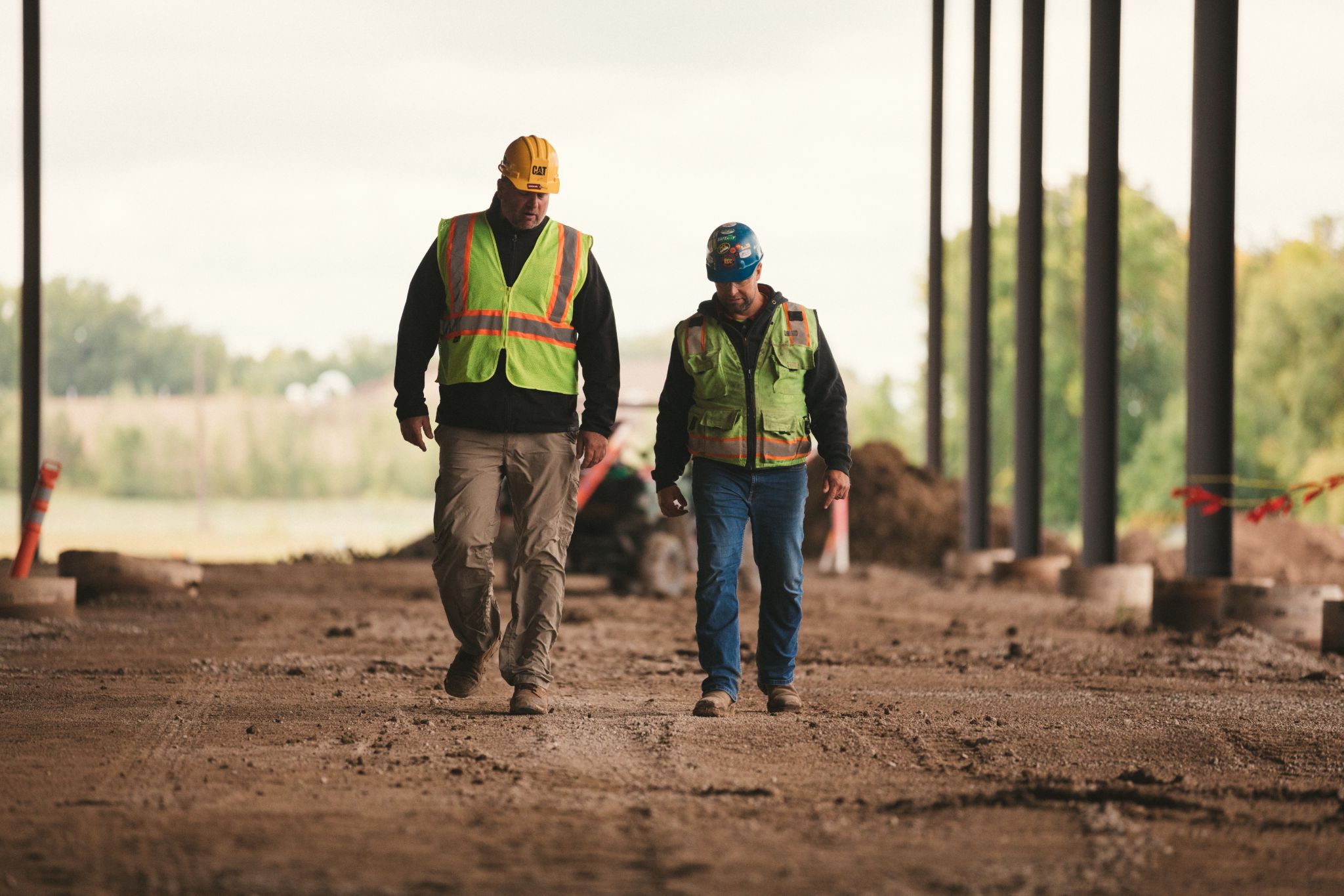 Two construction workers wearing safety vests and yellow and blue hardhats walking through a construction site
