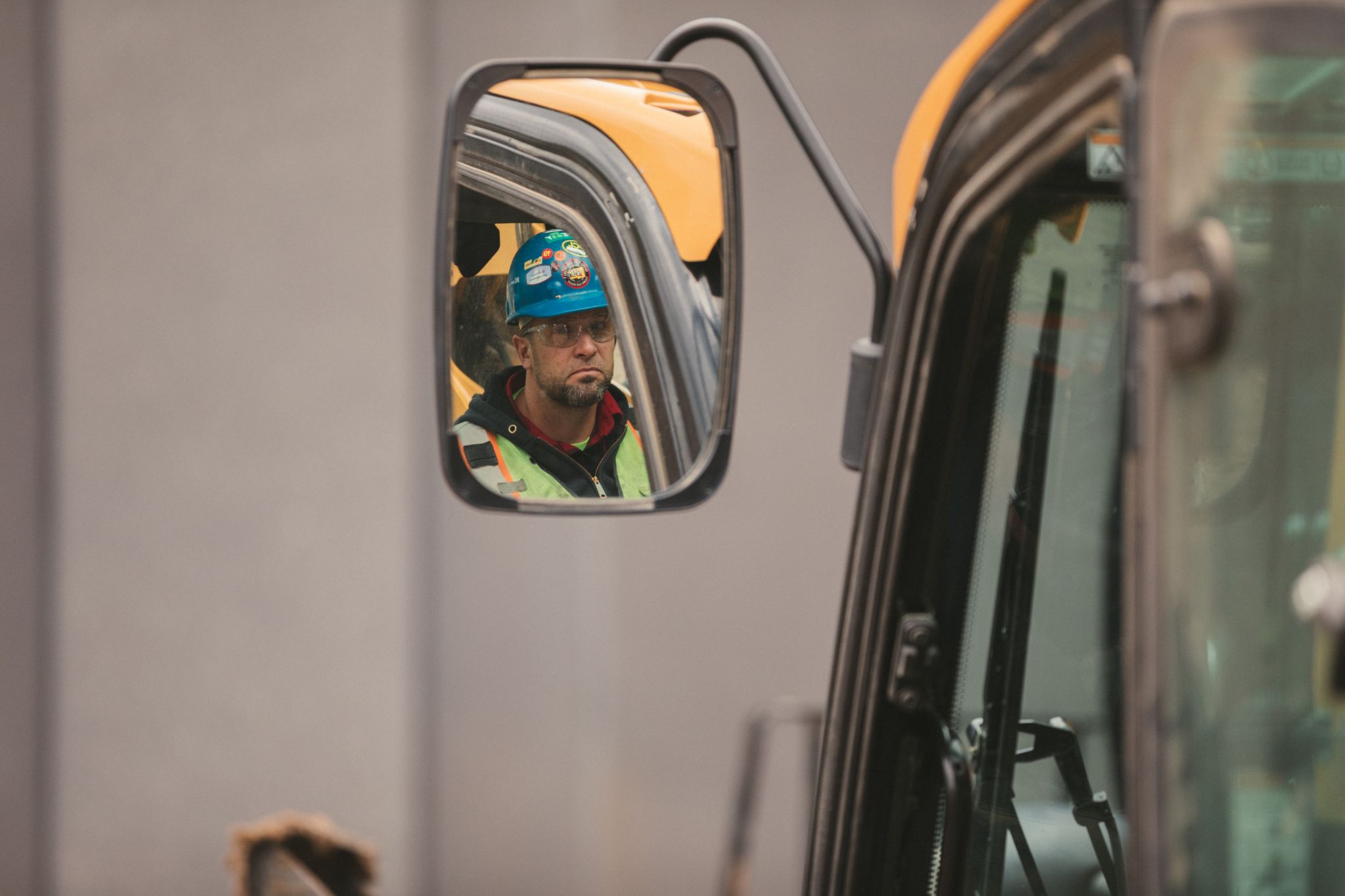 View of a construction worker wearing a blue hardhat and safety vest in side mirror of a construction vehicle