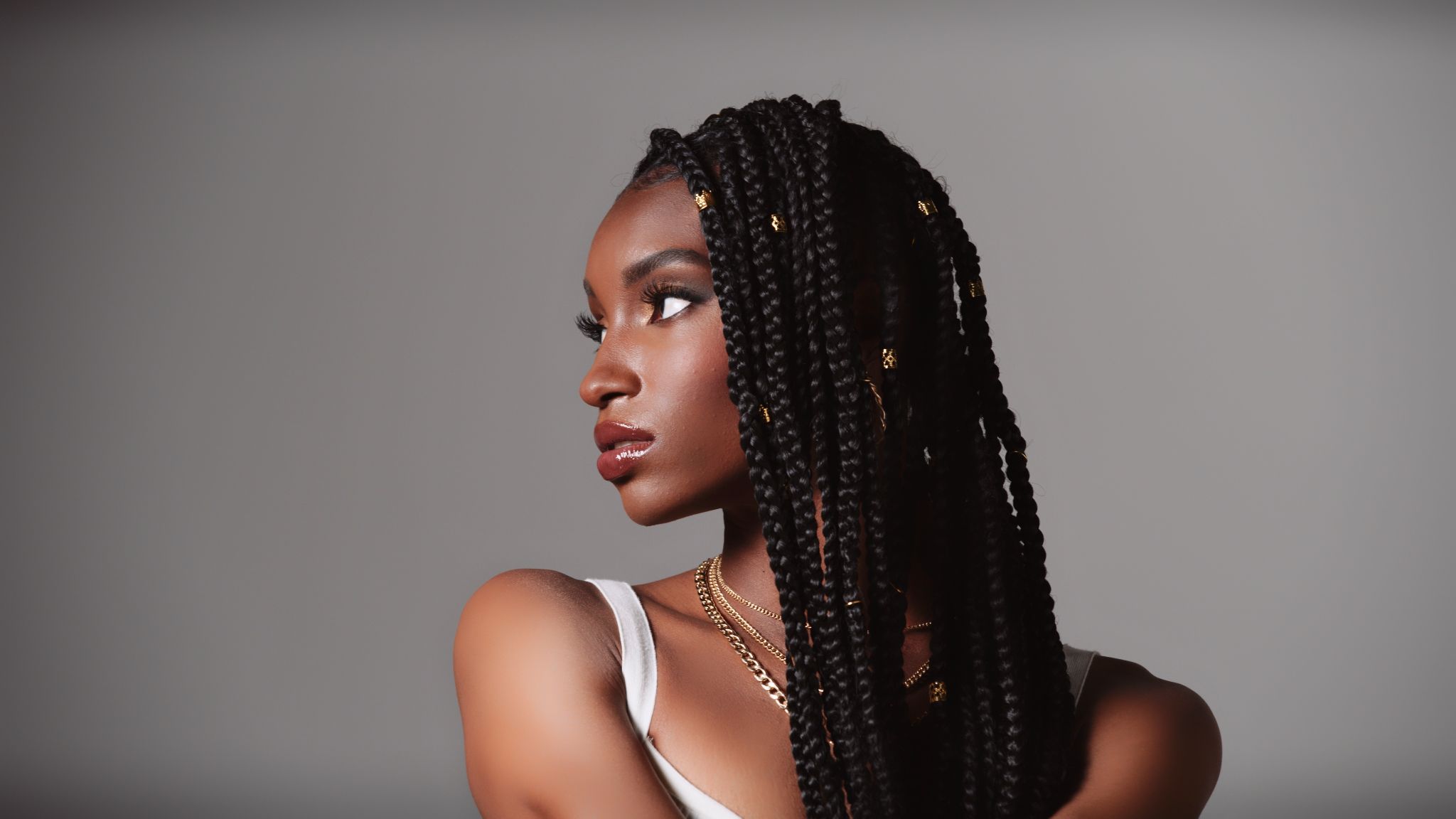 Female African American model headshot with black, braided hair wearing red lipstick, necklaces and white top posing for the camera looking off to the side