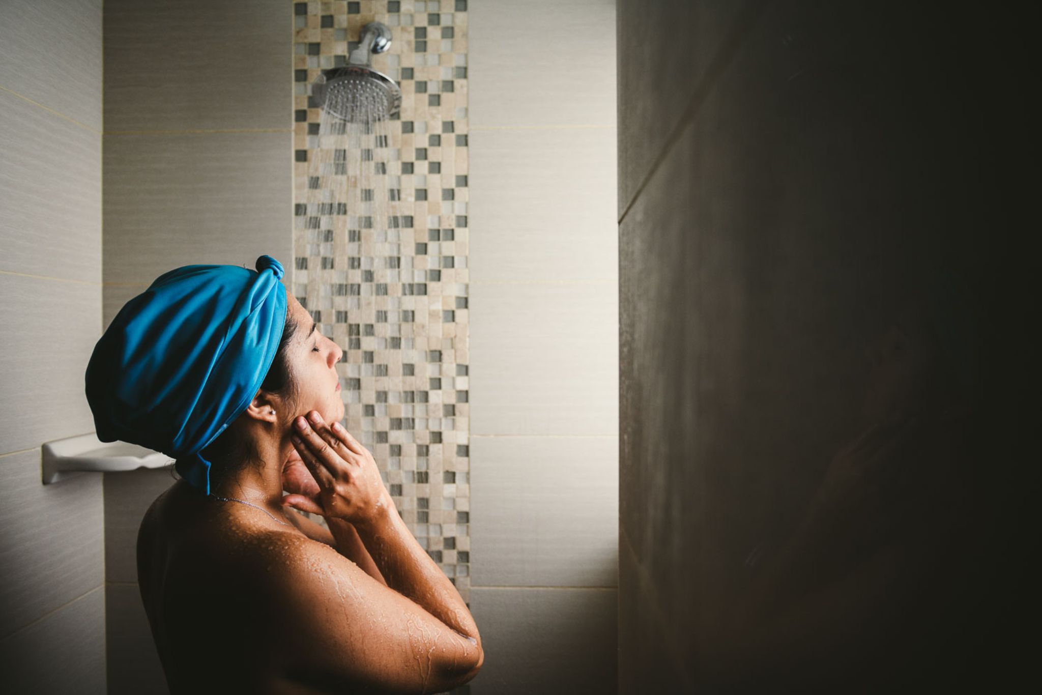 Side profile of woman wearing blue hair towel in the shower with eyes closed and water falling on her